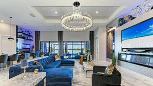 Ein modernes Wohnzimmer mit einem großen Ecksofa, einer Kristallleuchtre, einem Flachbildfernseher und einer Bar mit Regalen und Dekoration.