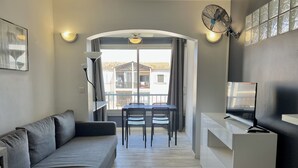 A modern living room with a grey sofa, a dining table with chairs, a TV, and a balcony with a view of buildings. - Bright 26 m² with pool in Frejus (Fréjus)