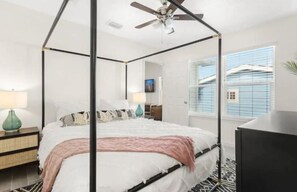 Un lit à baldaquin avec un ventilateur de plafond, une table de chevet avec une lampe, et une vue sur l’extérieur par une fenêtre munie de stores.