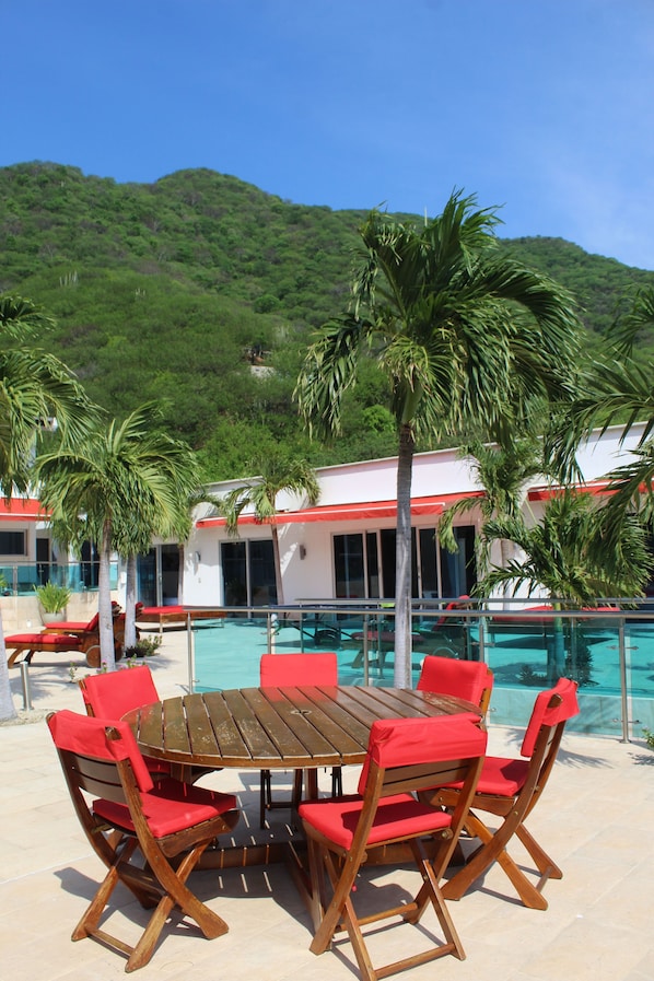 A poolside area with a wooden table and red chairs, surrounded by palm trees and a mountain backdrop.