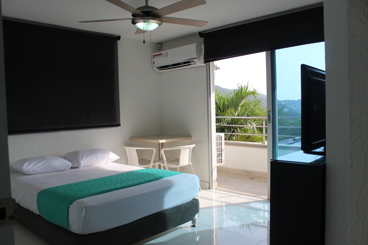 A bedroom with a bed, a ceiling fan, an air conditioning unit, a small table, and a balcony with a view of greenery and hills.