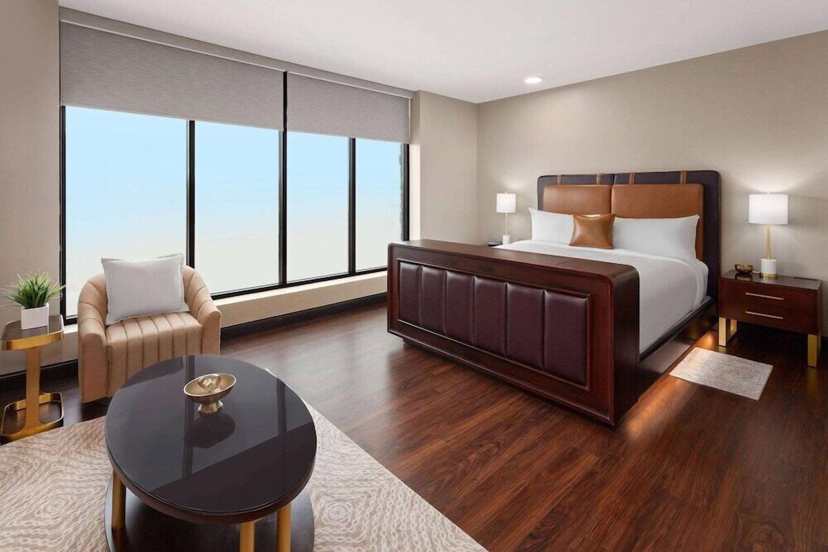 Een hotelkamer met een groot raam, een bed, een stoel, een tafel en een nachtkastje.