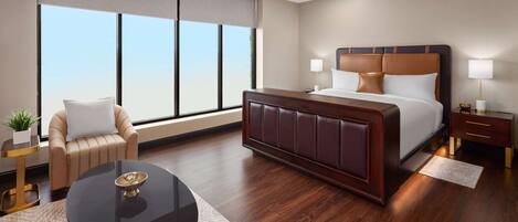 A modern hotel room with a large bed, a round coffee table, a sofa, and a view of the sky through large windows.