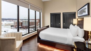 A hotel room with a large bed, a chair, a desk, and a view of a cityscape through large windows. - Legacy Hotel Trademark Collection By Wyndham (Green Bay)