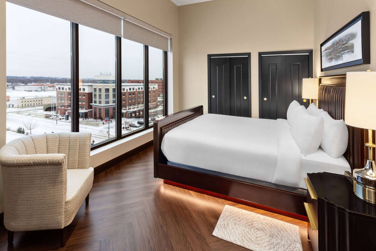 Hotelkamer met een groot bed, een stoel, een bureau en uitzicht op een stadslandschap door grote ramen.