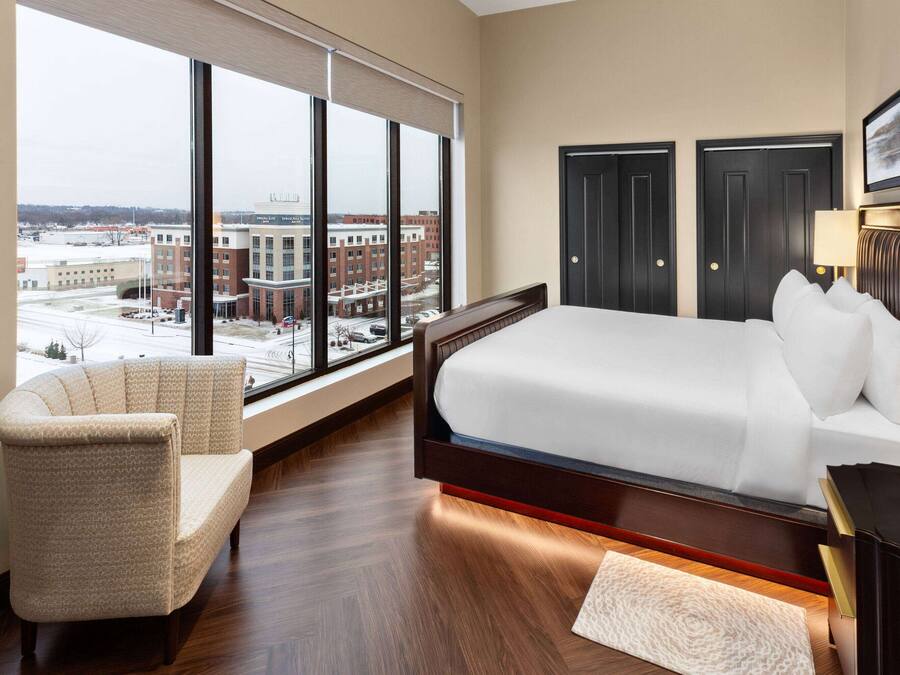 Quarto de hotel com uma cama grande, uma cadeira, uma escrivaninha e vista para a paisagem urbana através de janelas amplas.