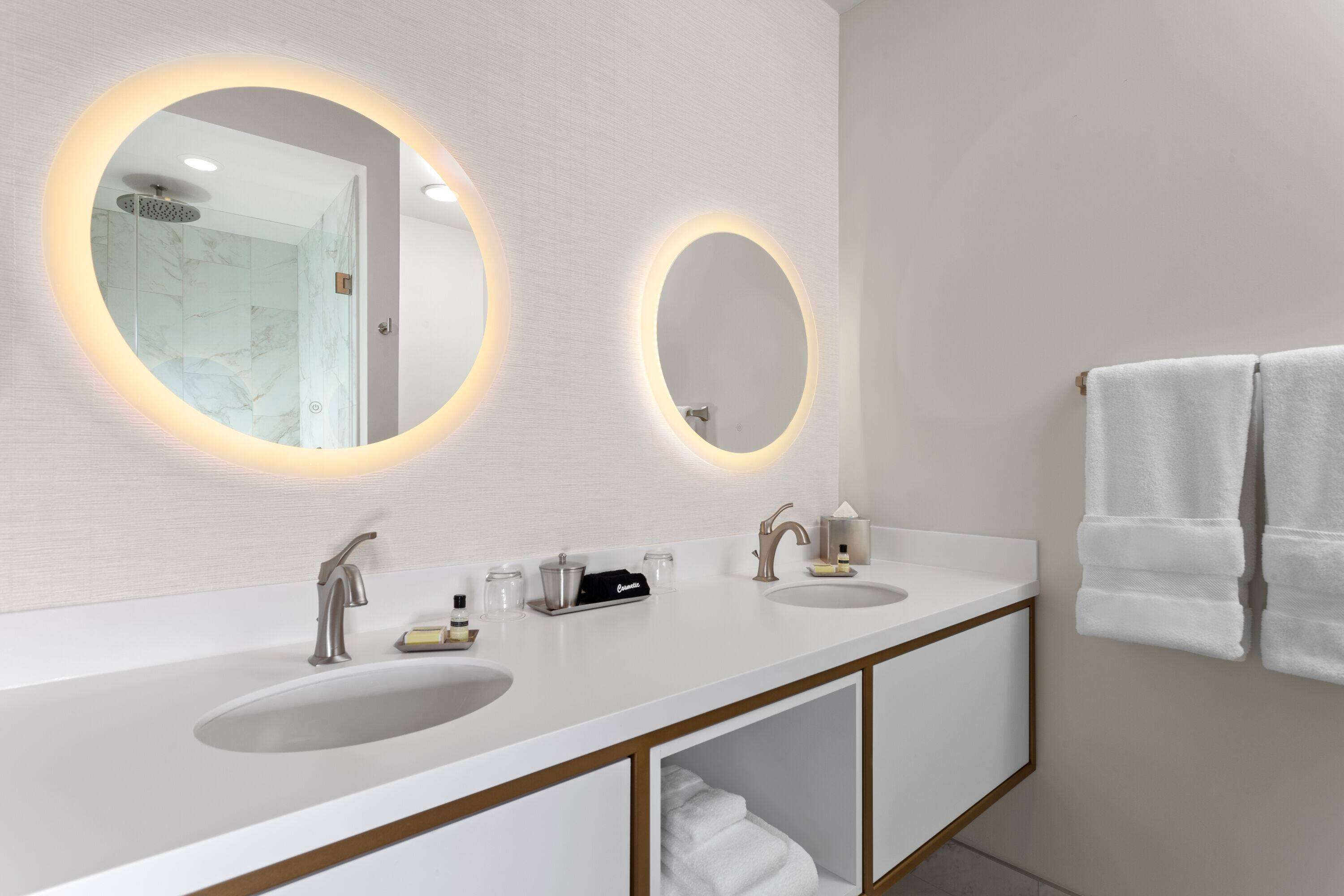 Un baño moderno con dos lavamanos, un espejo grande con iluminación perimetral y un perchero con toallas dobladas.