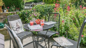A patio with metal chairs and a table, surrounded by lush greenery and flowers. - 2 bedroom accommodation in Eyam, near Bakewell (Eyam, near Bakewell)