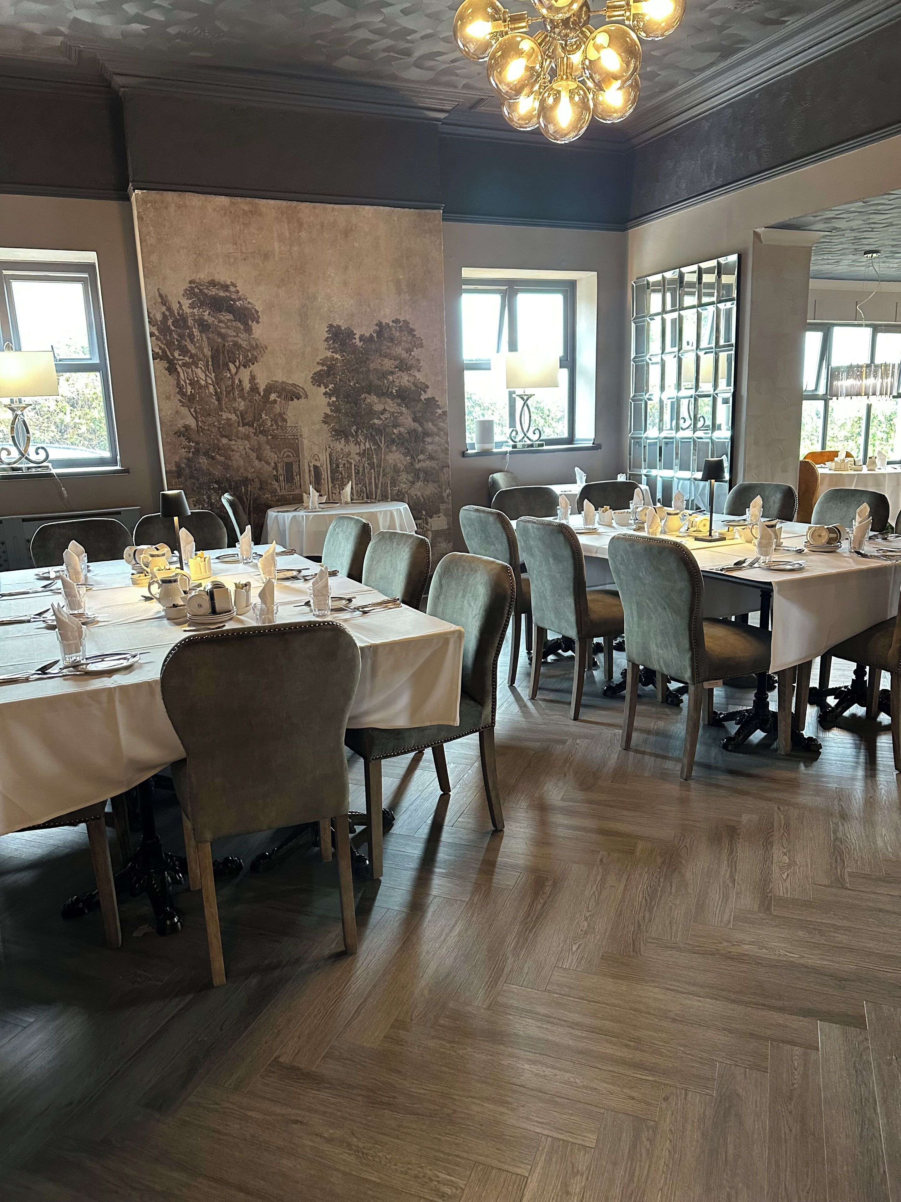 A restaurant with multiple tables set for dining, featuring a herringbone wood floor and a large mural on the wall.