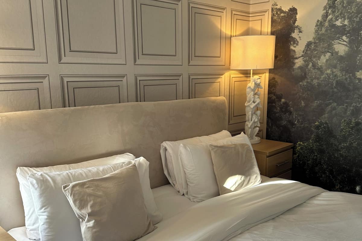 A neatly made bed with white linens and pillows, a wall with a geometric pattern, and a lamp on a bedside table.