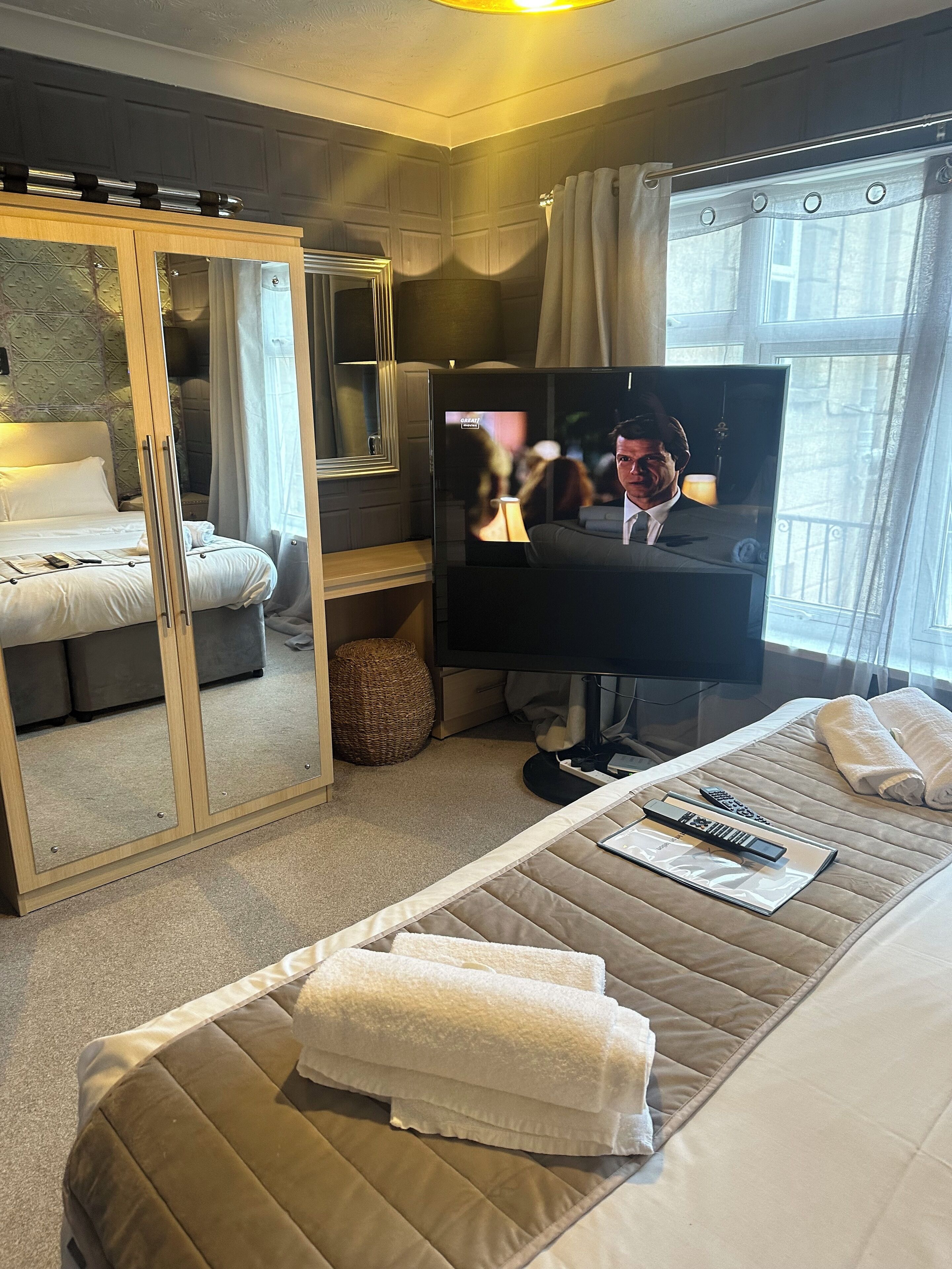 A hotel room with a bed, a television, a mirror, and a window with curtains.
