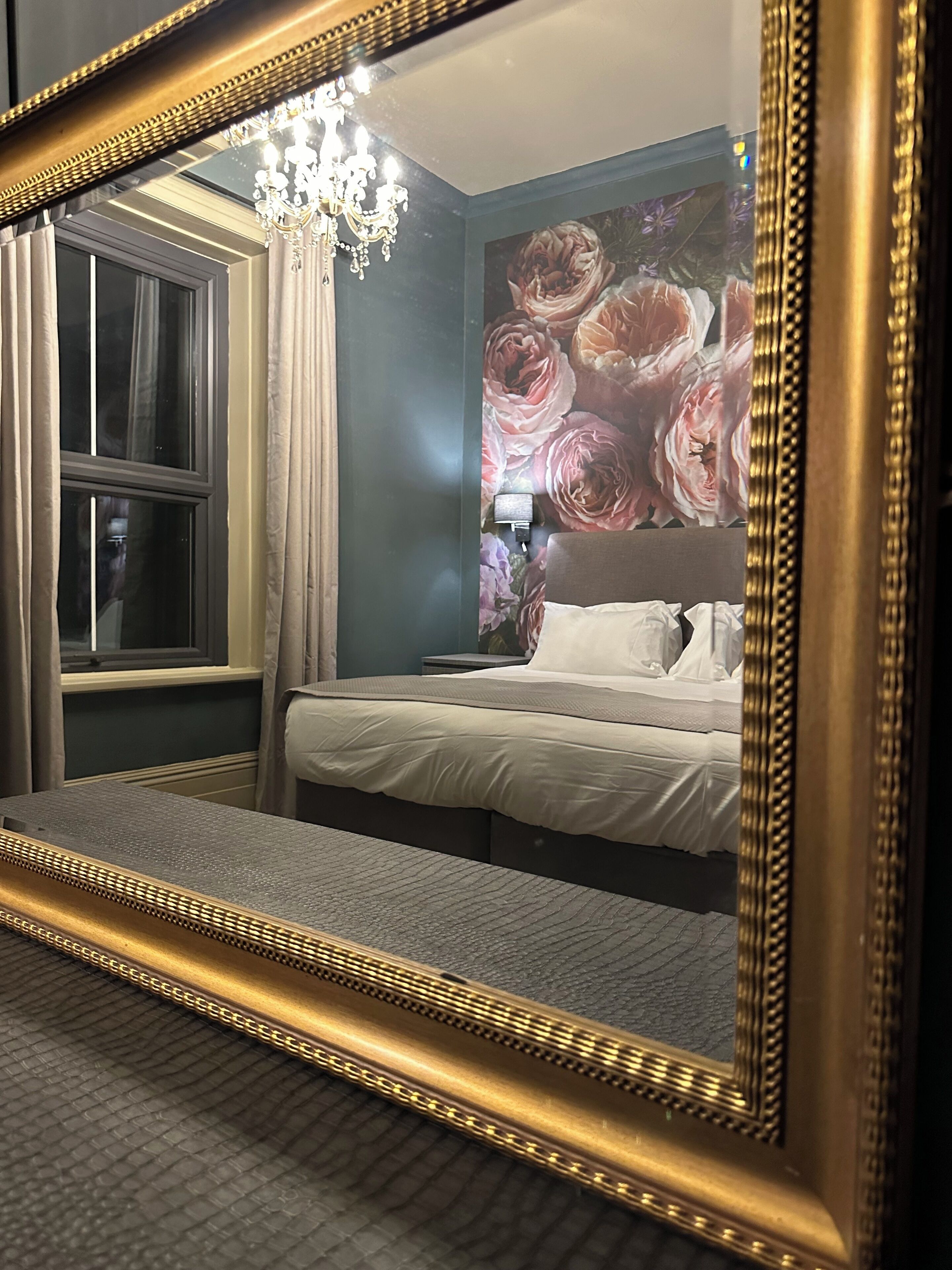 A hotel room with a large mirror, a bed with white linens, a chandelier, and a floral wallpaper.