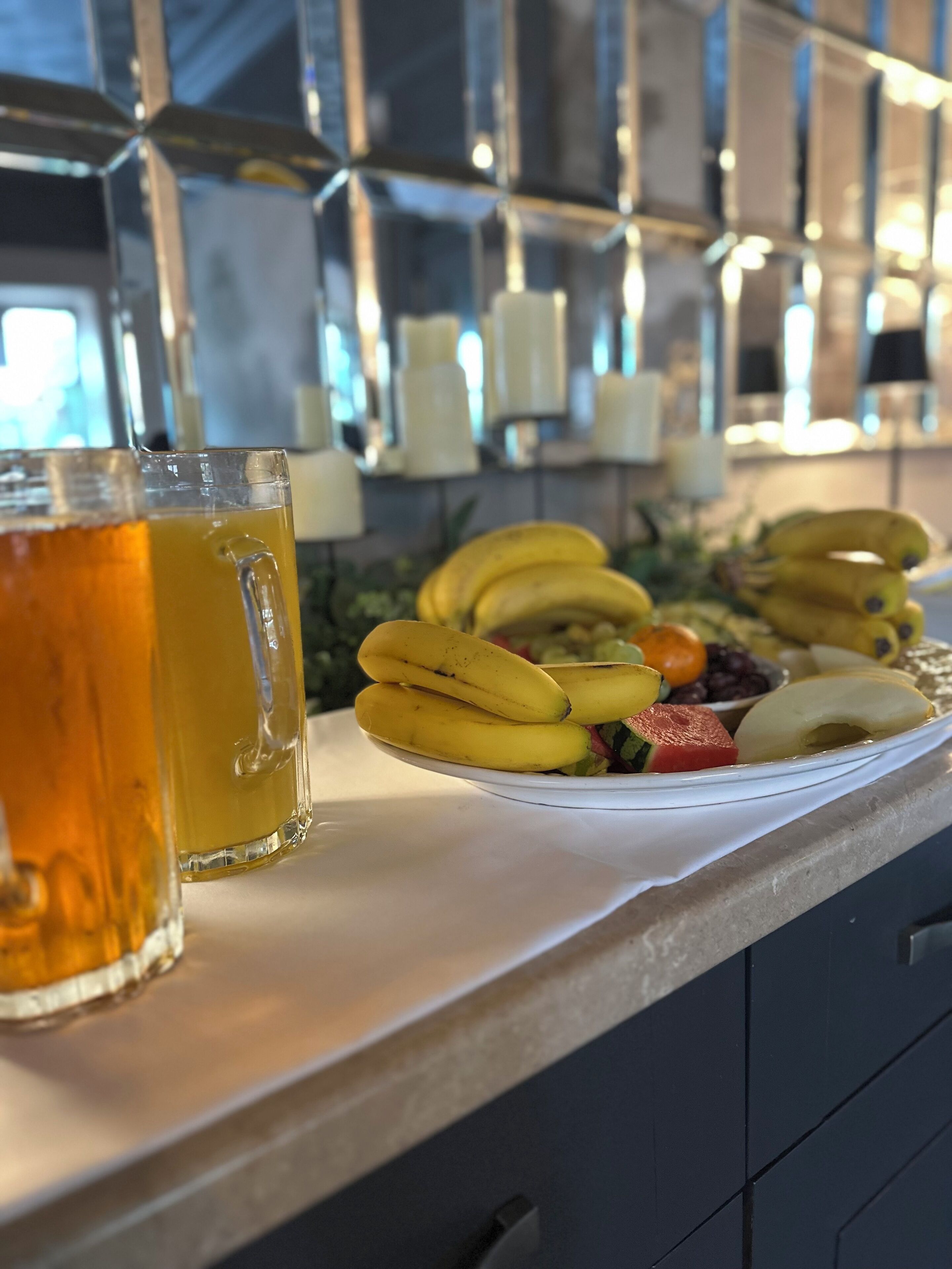 A counter with a glass of amber-colored beverage, a plate of bananas, and various fruits.