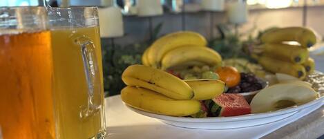 A counter with a glass of amber-colored beverage, a plate of bananas, and various fruits.