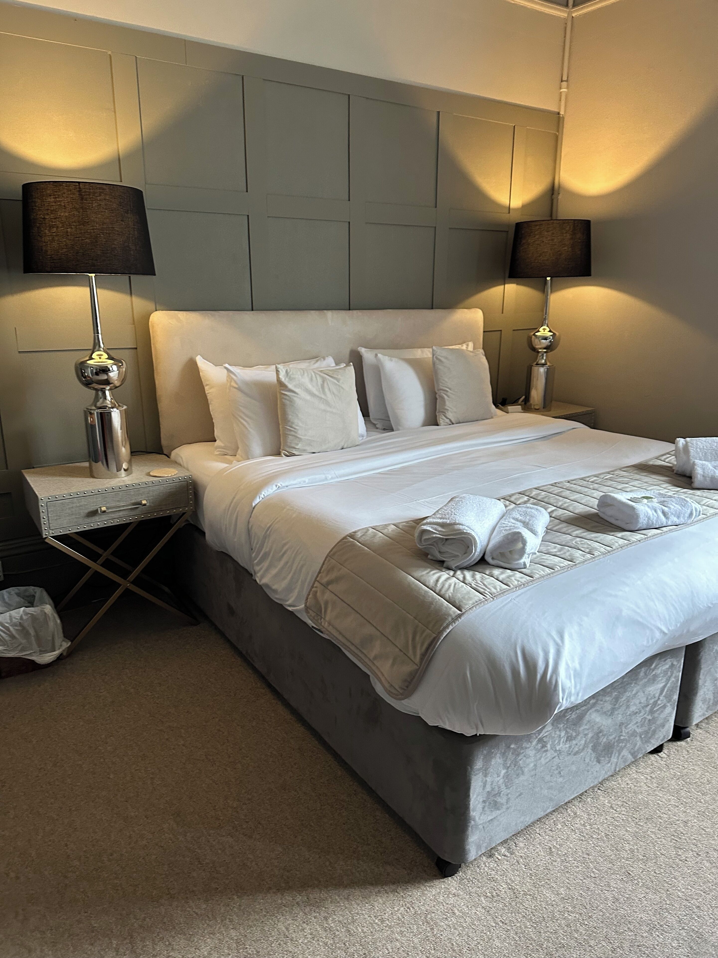 A neatly made bed with white linens and pillows, two bedside lamps, a small bedside table, and a trash bin.