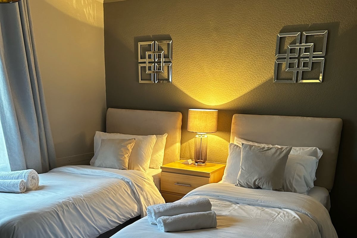 A hotel room with two beds, a nightstand with a lamp, and a wall-mounted decorative piece.