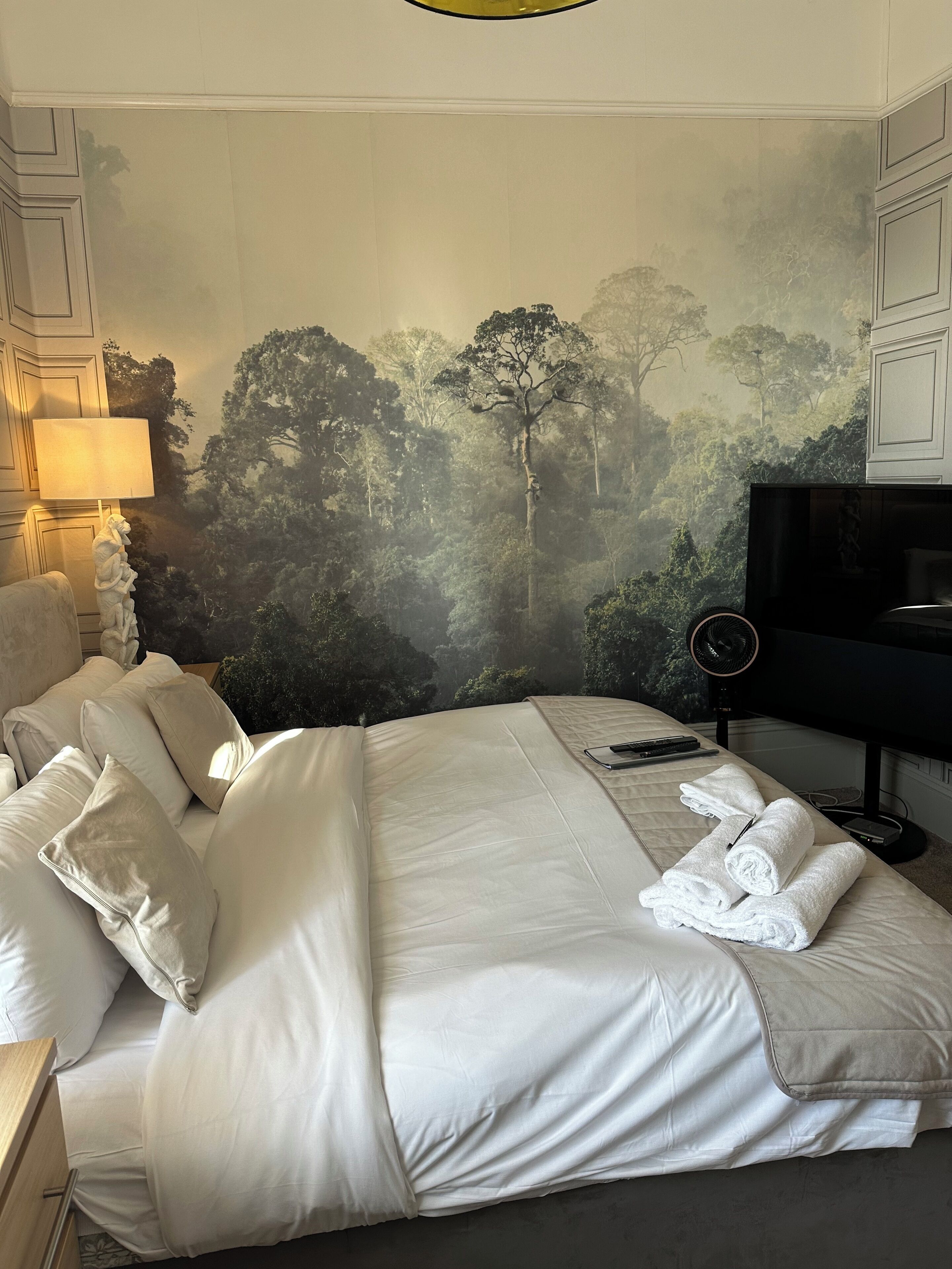 Un dormitorio con una cama grande, ropa de cama blanca, un televisor de pantalla plana, un ventilador de pared y un cuadro de un paisaje boscoso.