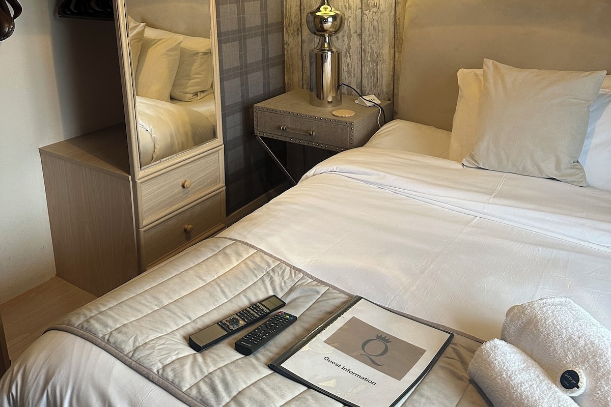 A hotel room with a bed, a bedside table with a lamp, a bedside table with a remote control, a bedside table with a book, and a wardrobe.