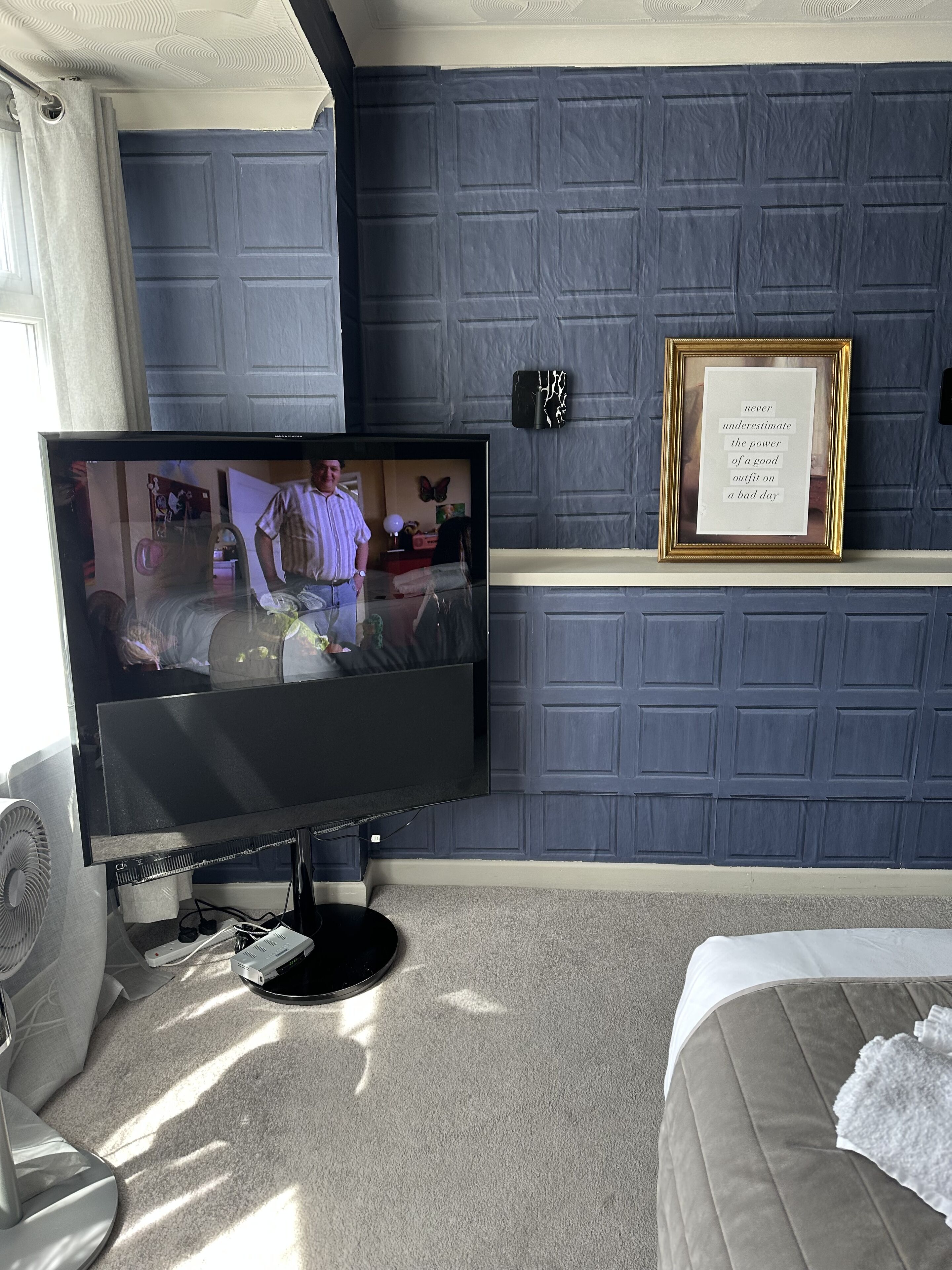 Una habitación con una televisión sobre un mueble bajo, una cama con un almohárraga blanca y una pared decorada con un marco con una cita.