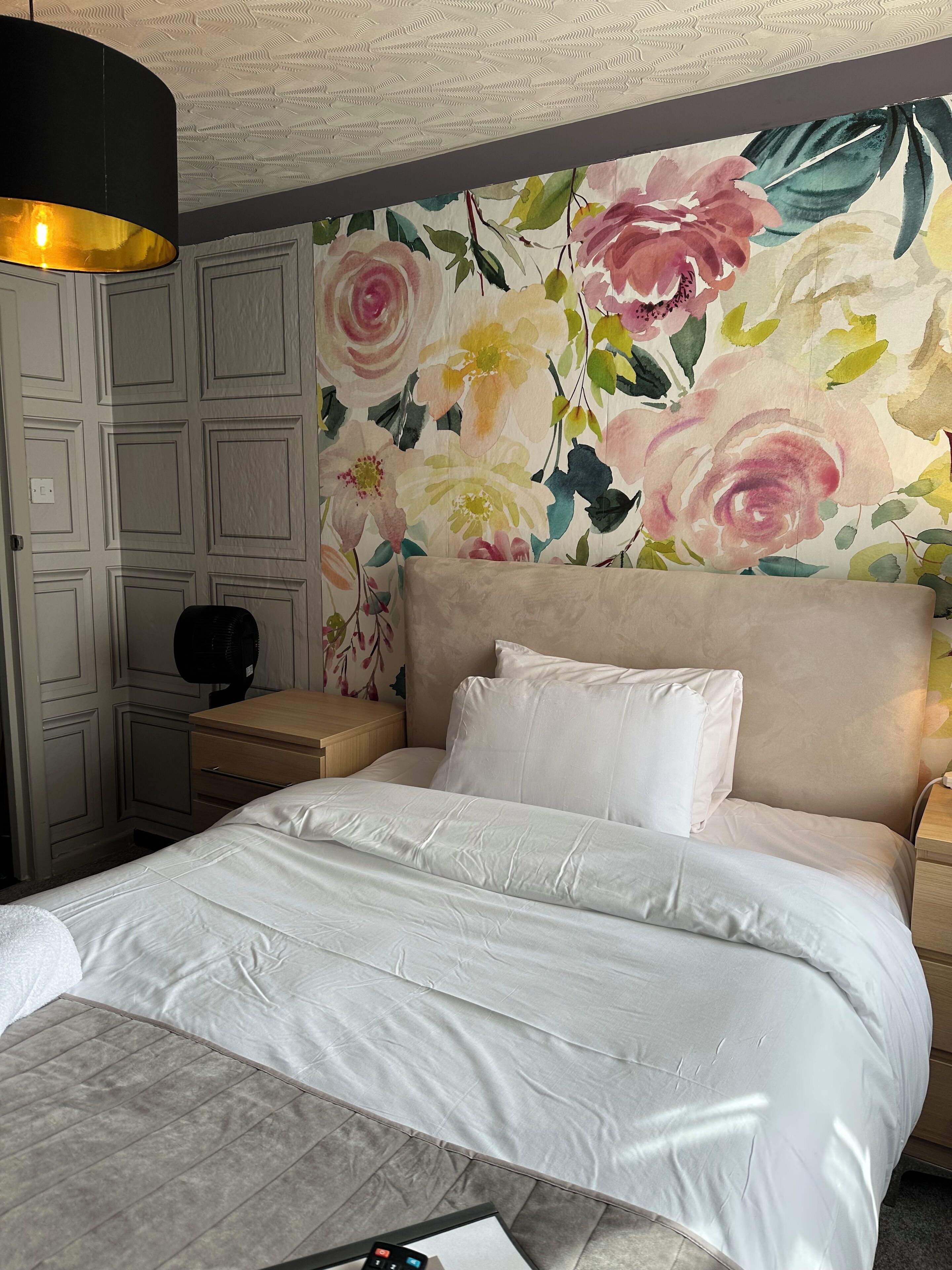 Un lit bien fait, avec du linge de lit blanc, un papier peint à motifs floraux et un luminaire moderne au plafond.