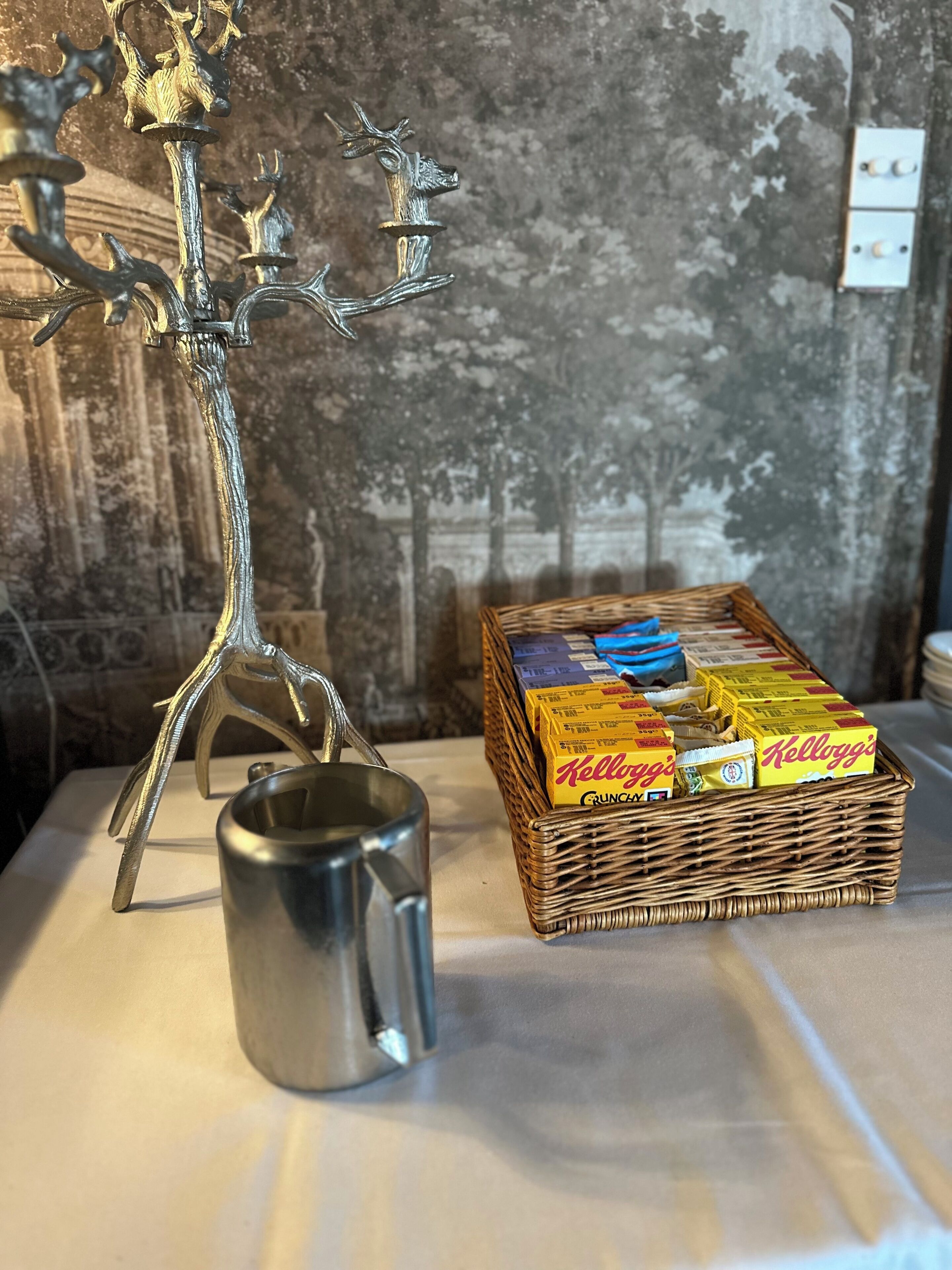 A metal candle holder with a tree design, a metal mug, and a wicker basket containing Kellogg's cereal boxes.