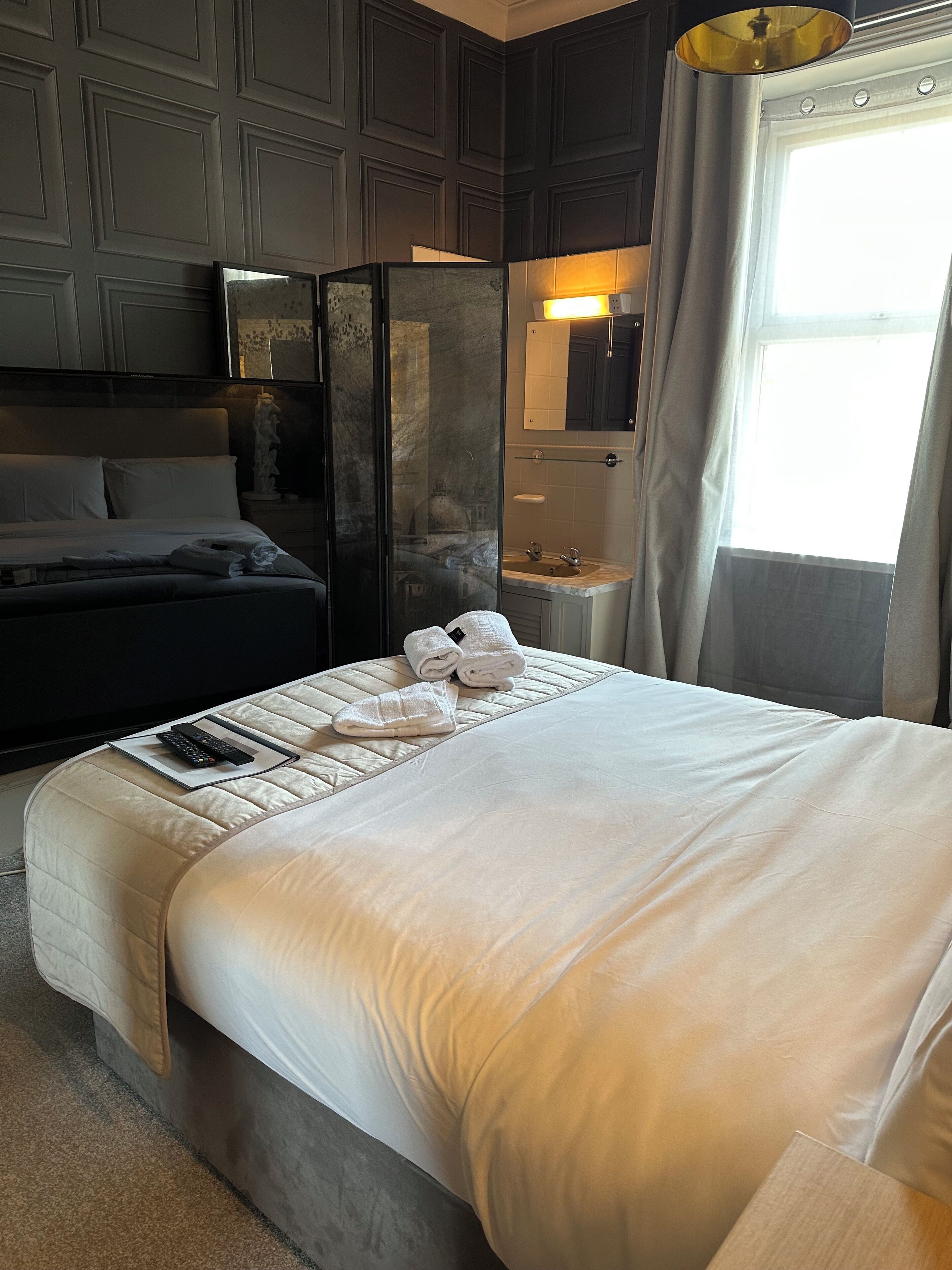 A hotel room with a large bed, a nightstand with a lamp, a bathroom with a mirror, and a view of the outside.