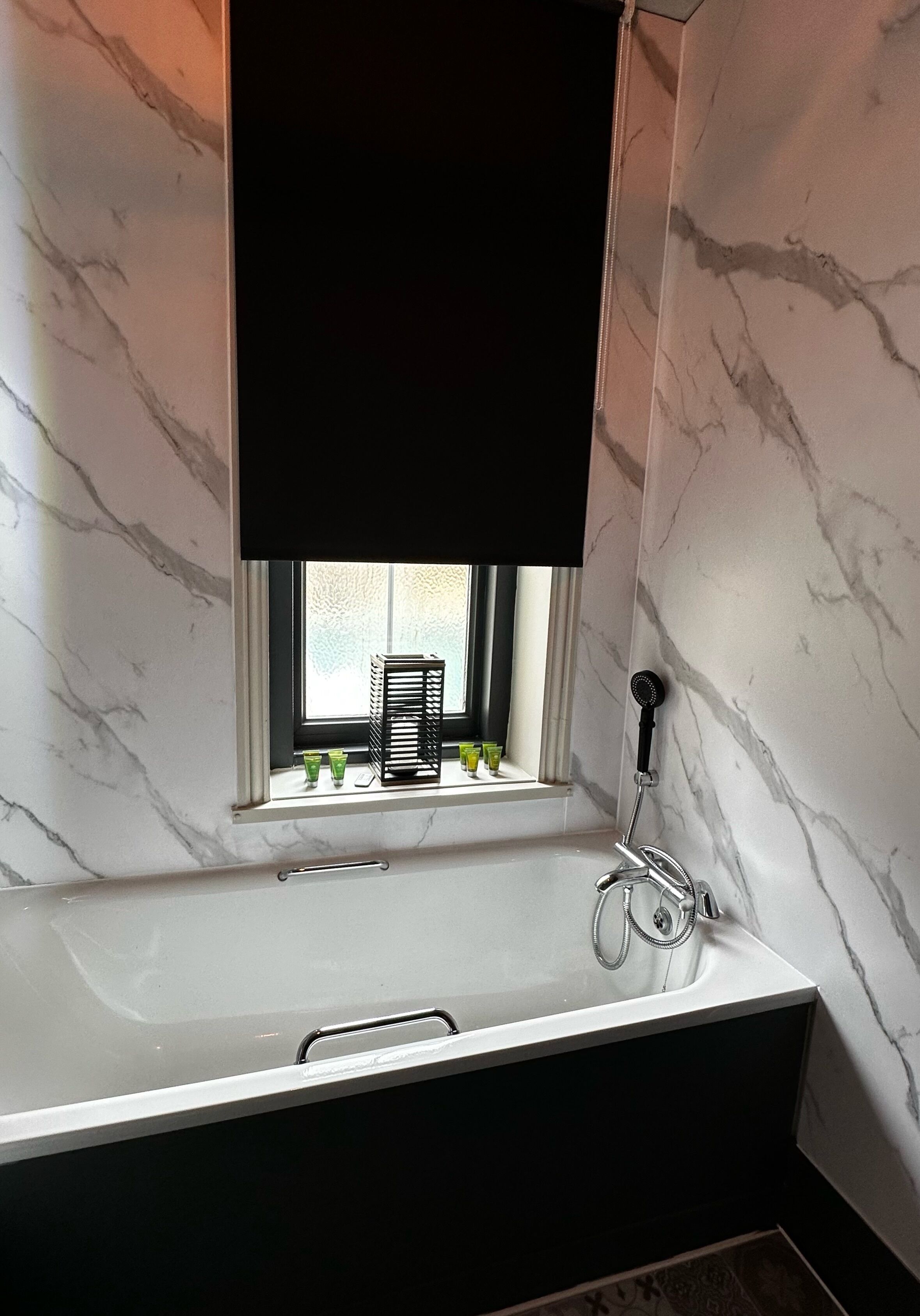 A bathroom with a marble wall, a window with a black blind, a shower, and a small shelf with toiletries.