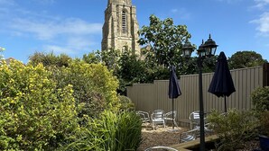 An outdoor patio with metal chairs and a table, surrounded by greenery and a tall church spire in the background. - The Q Hotel (Shanklin)