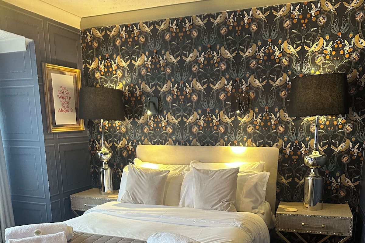 A neatly made bed with white linens, a bedside table with a lamp, and a wall with a floral pattern.