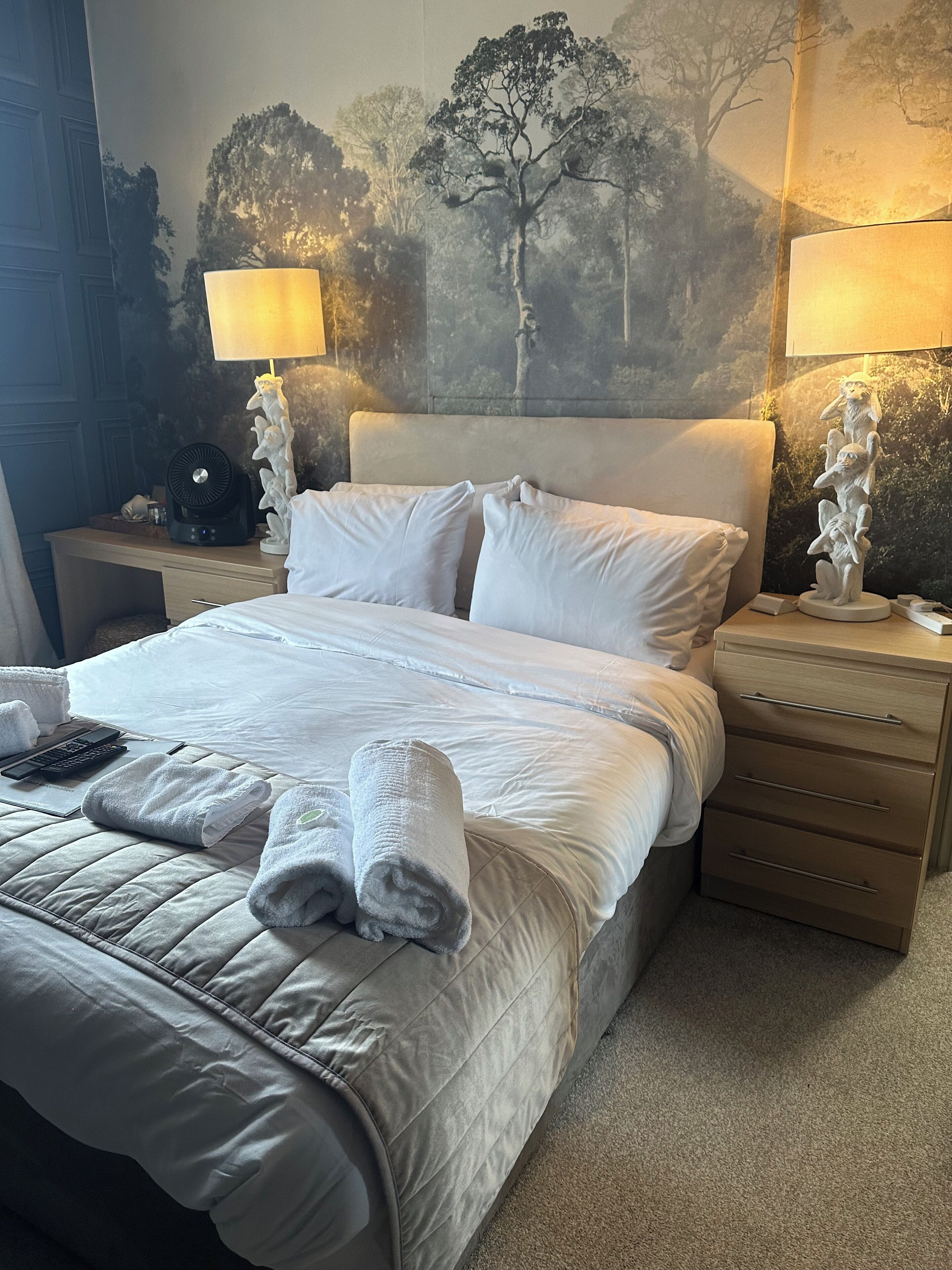 A neatly made bed with white linens and towels, a bedside table with a lamp, and a wall mural of trees.