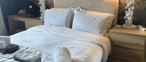 A neatly made bed with white linens and towels, a bedside table with a lamp, and a wall mural of trees.