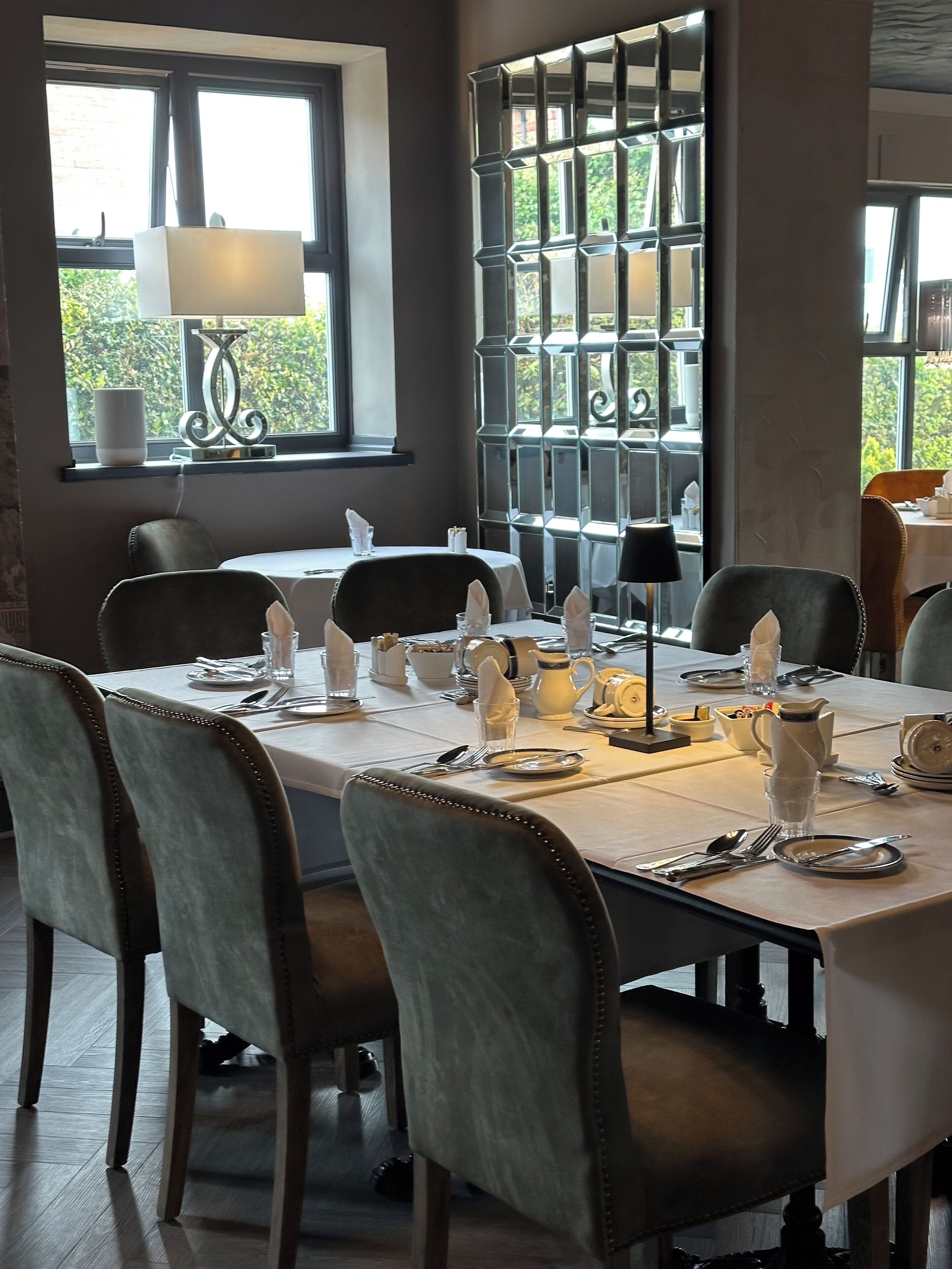 A dining area with a table set for four, including plates, cutlery, and glasses. There are chairs around the table, and a window with a view of greenery outside.