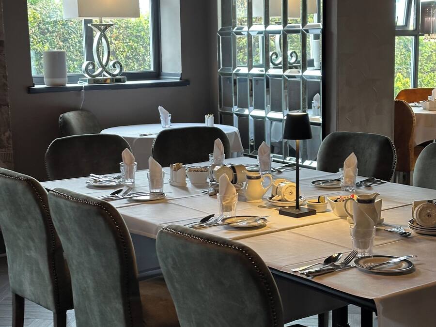 A dining area with a table set for four, including plates, cutlery, and glasses. There are chairs around the table, and a window with a view of greenery outside.