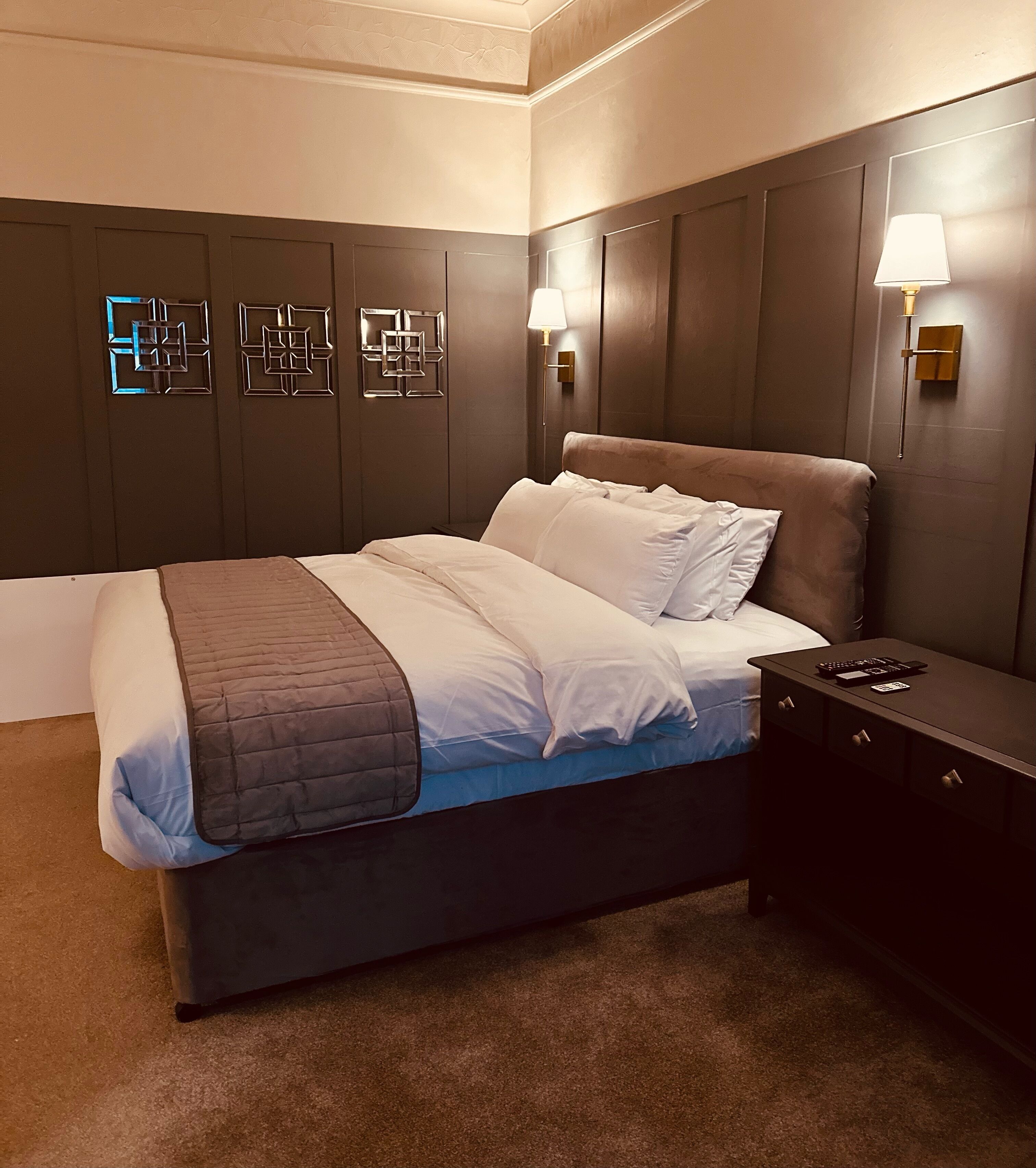 A modern bedroom with a dark brown headboard, a large bed with white linens, a bedside table with a lamp, and geometric wall art.