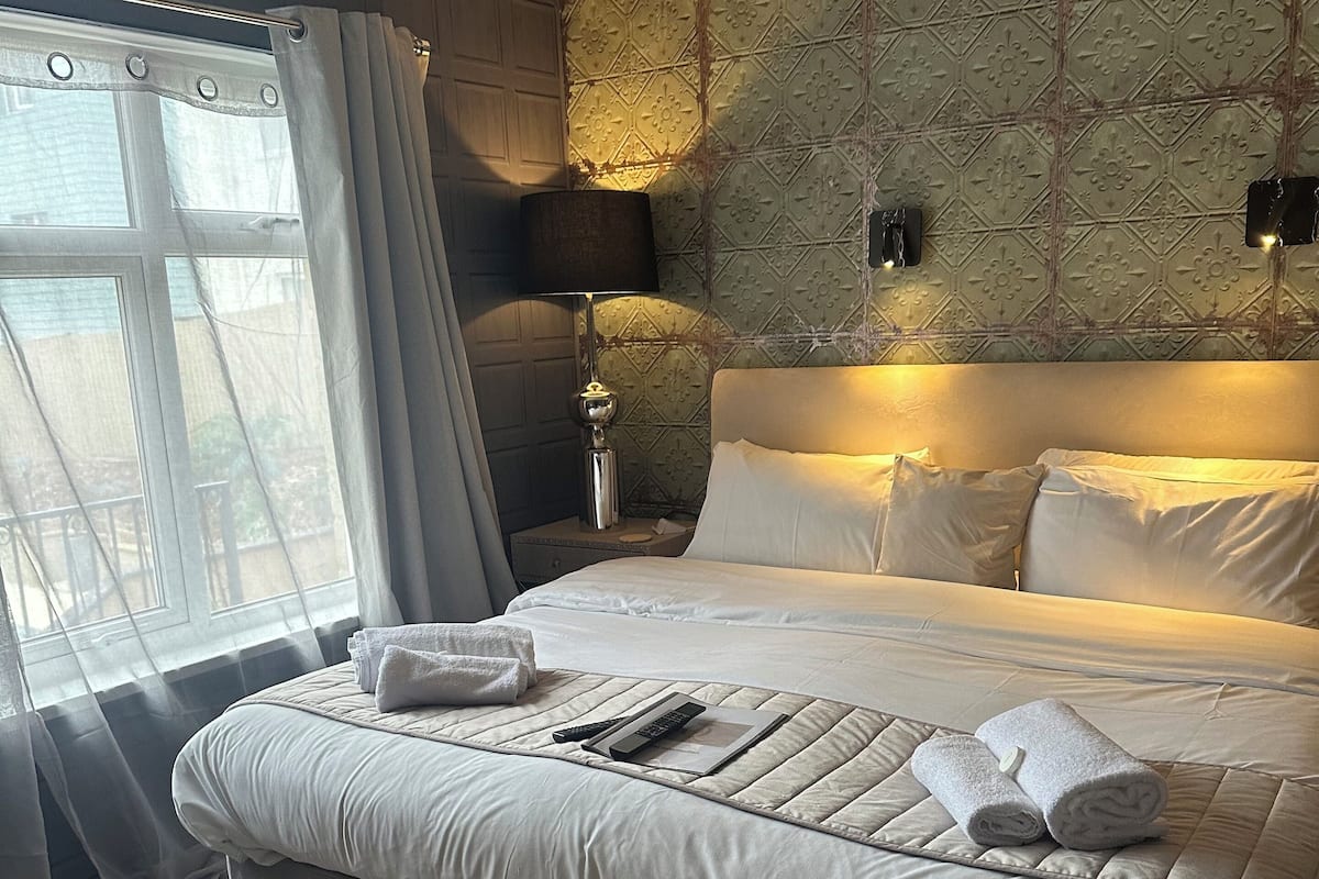 A large bed with a quilted headboard, a bedside table with a lamp, and a window with sheer curtains.