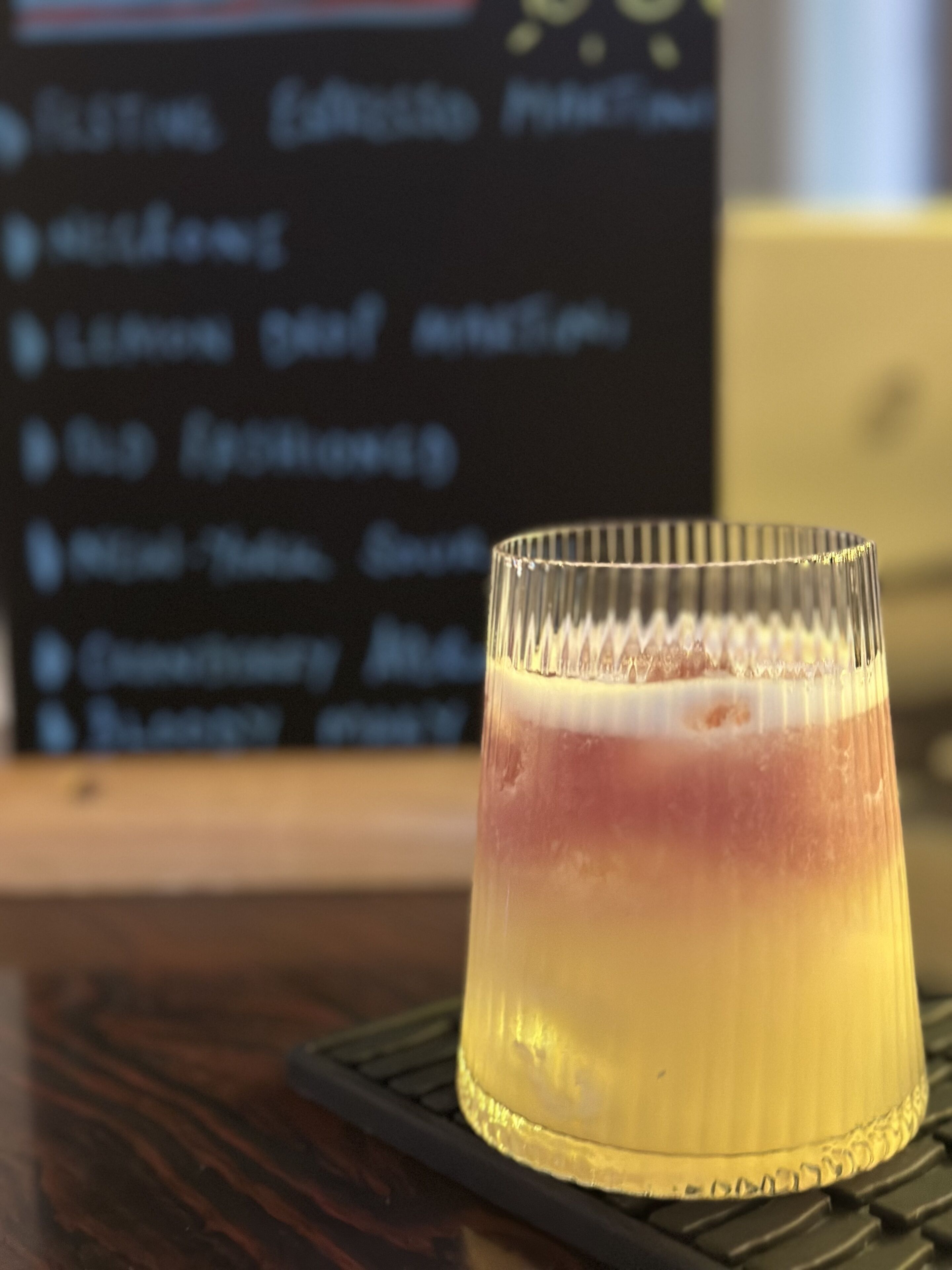 A cocktail glass with a layered drink on a coaster, placed on a wooden surface with a blurred background.