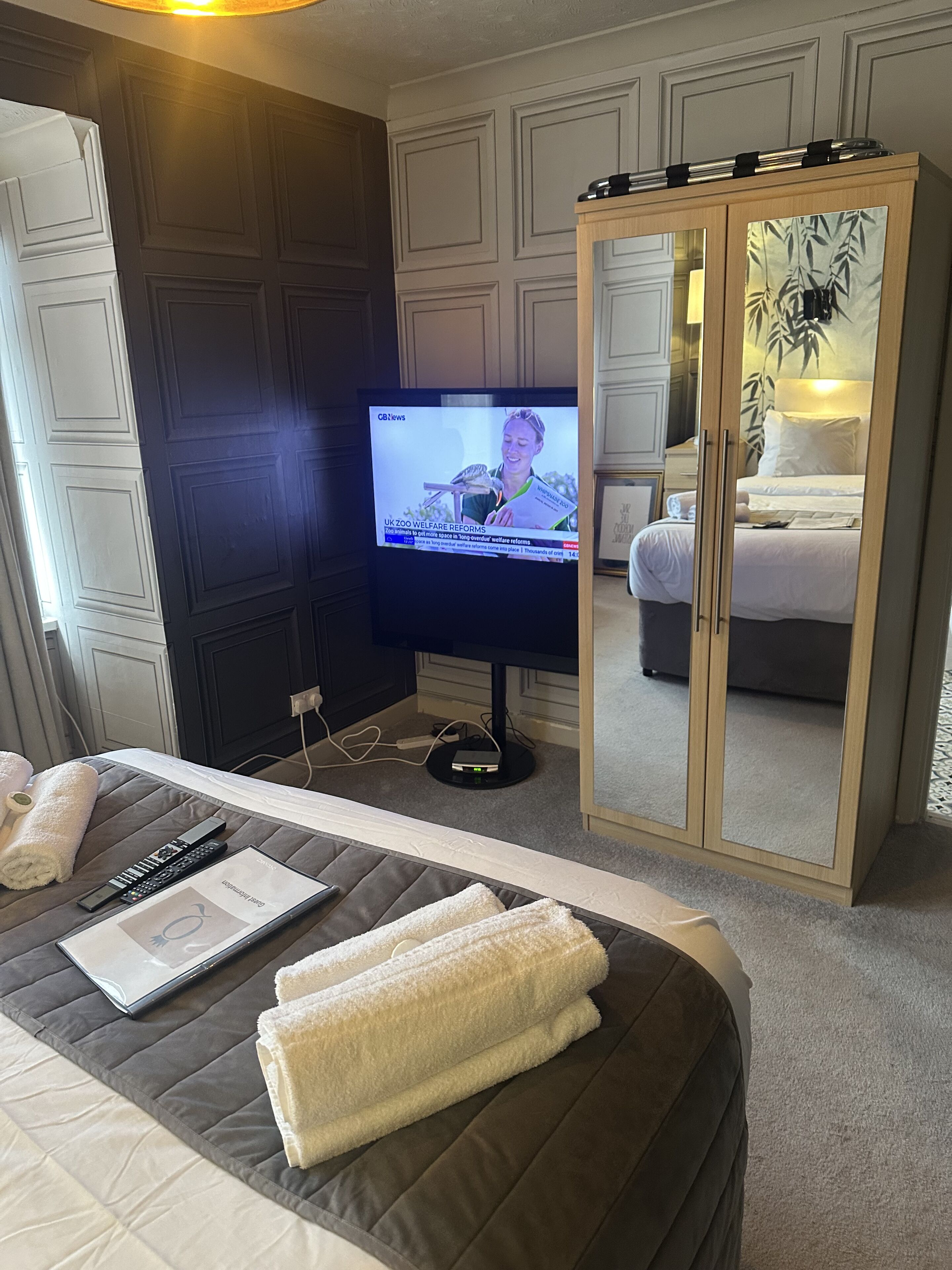 A hotel room with a bed, a TV displaying a news channel, a bedside table with a remote control and a magazine, and a wardrobe with a mirror.