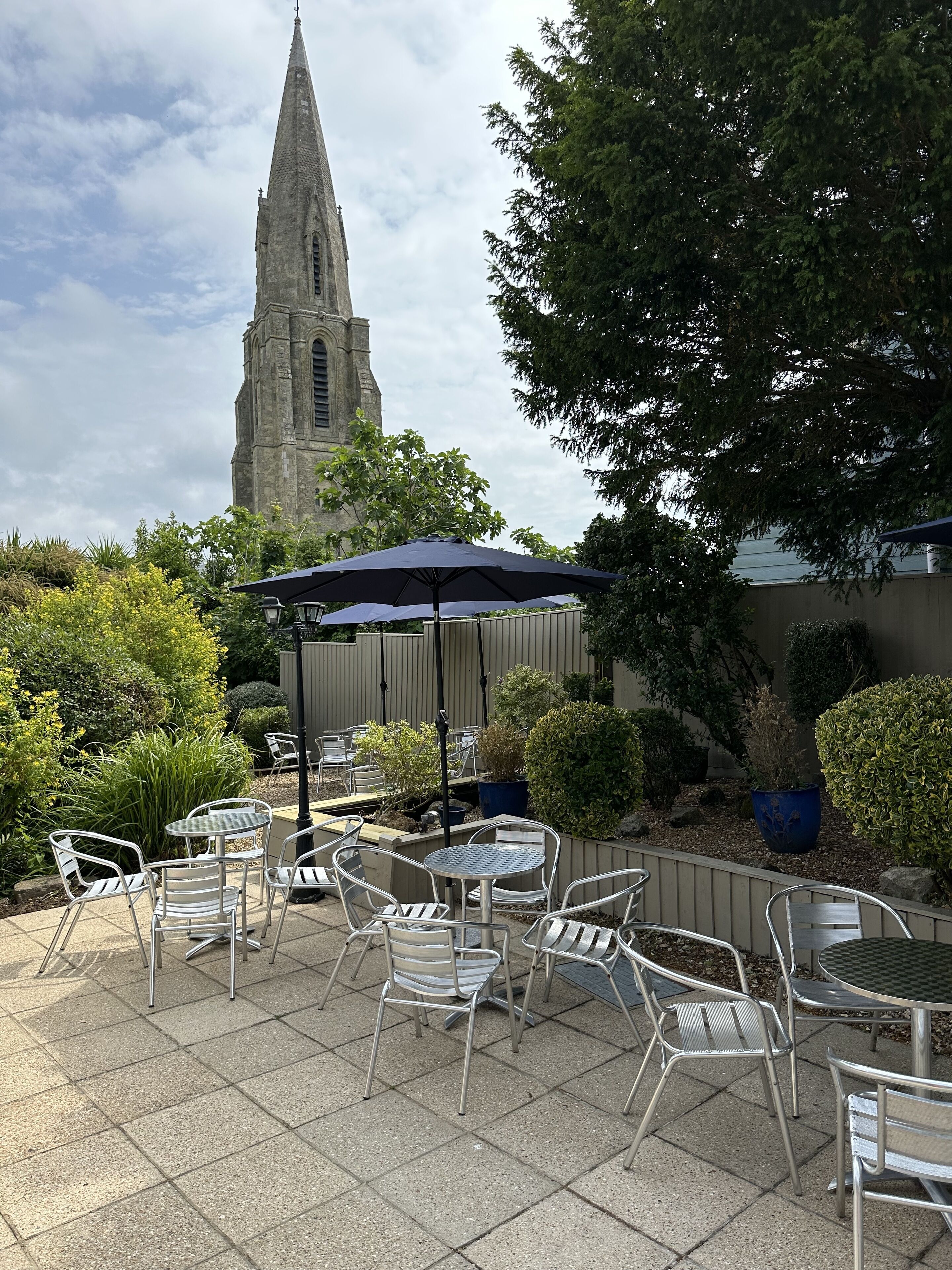 An outdoor patio with metal chairs and tables, a large umbrella, and a church tower in the background.