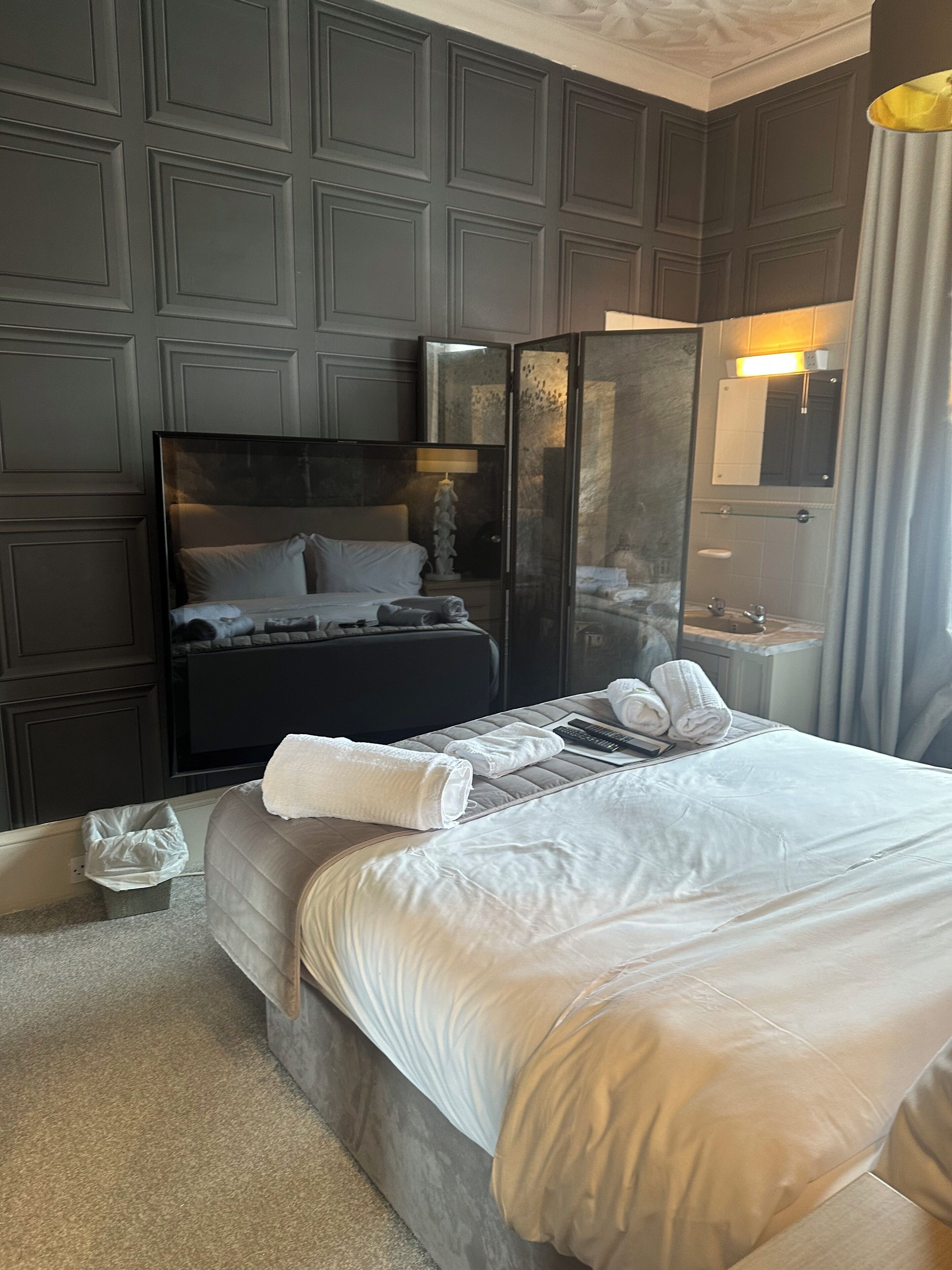 Quarto de hotel com uma cama grande, mesa de cabeceira, espelho e banheiro visível através de um painel de vidro.