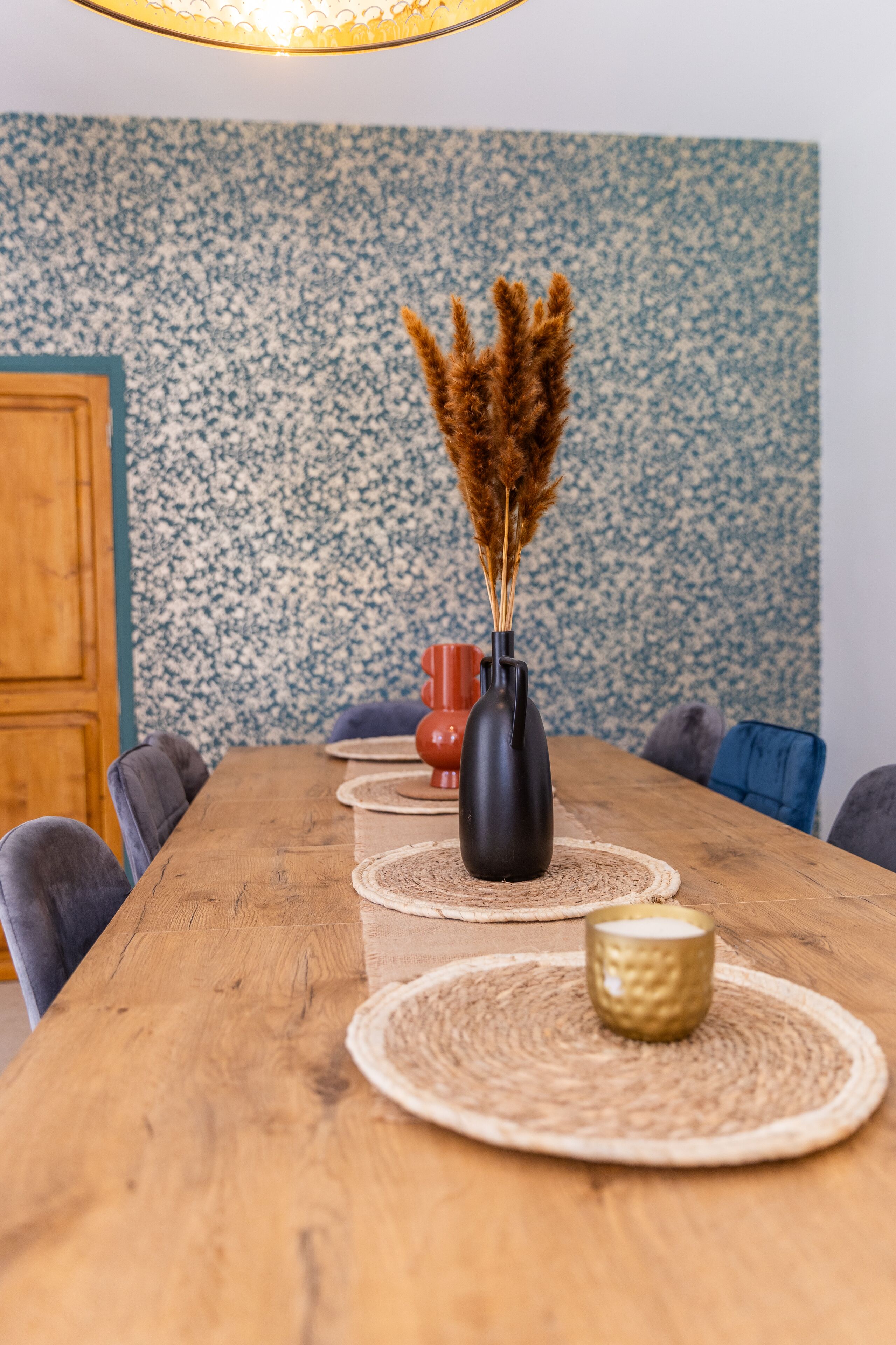 Una mesa de comedor de madera con un florero negro y pastos de pampa, rodeada de individuales tejidos y sillas.