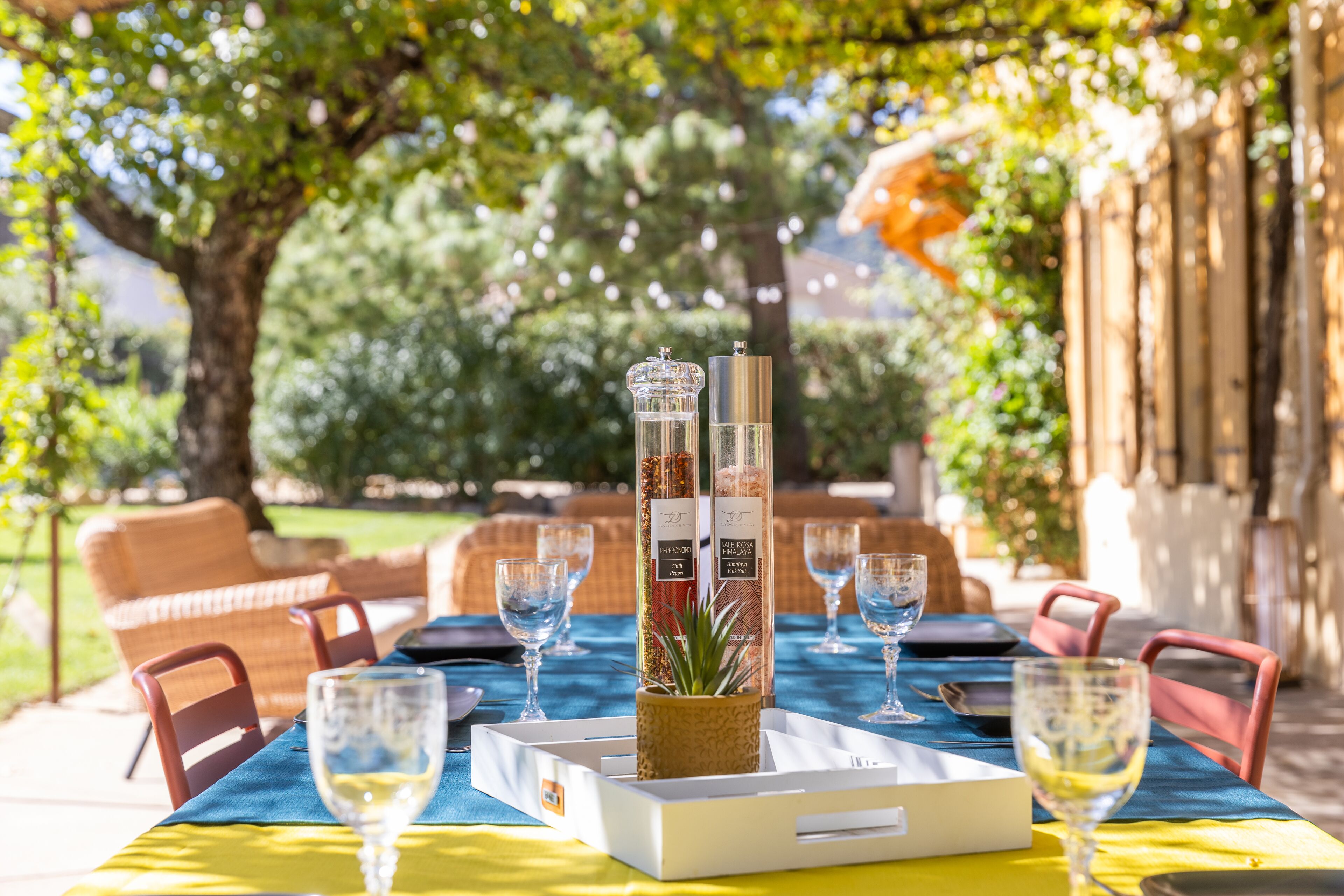 Un espace de restauration en plein air avec une table dressée pour deux personnes, agrémenté d’un centre de table en verre transparent, d’un présentoir à menus et de deux verres à vin.
