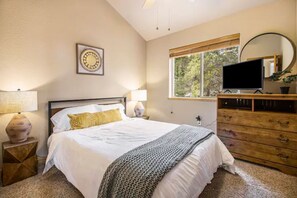 Une chambre à coucher comprenant un lit, des lampes de chevet, une commode, un téléviseur et une fenêtre donnant sur des arbres.