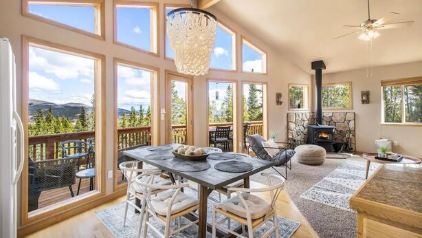 A spacious living room with a large dining table, comfortable seating, a fireplace, and a scenic mountain view through large windows. - *NEW* Stunning Mountain Views/Hot Tub/3BR/Sleeps 6 (Fairplay)