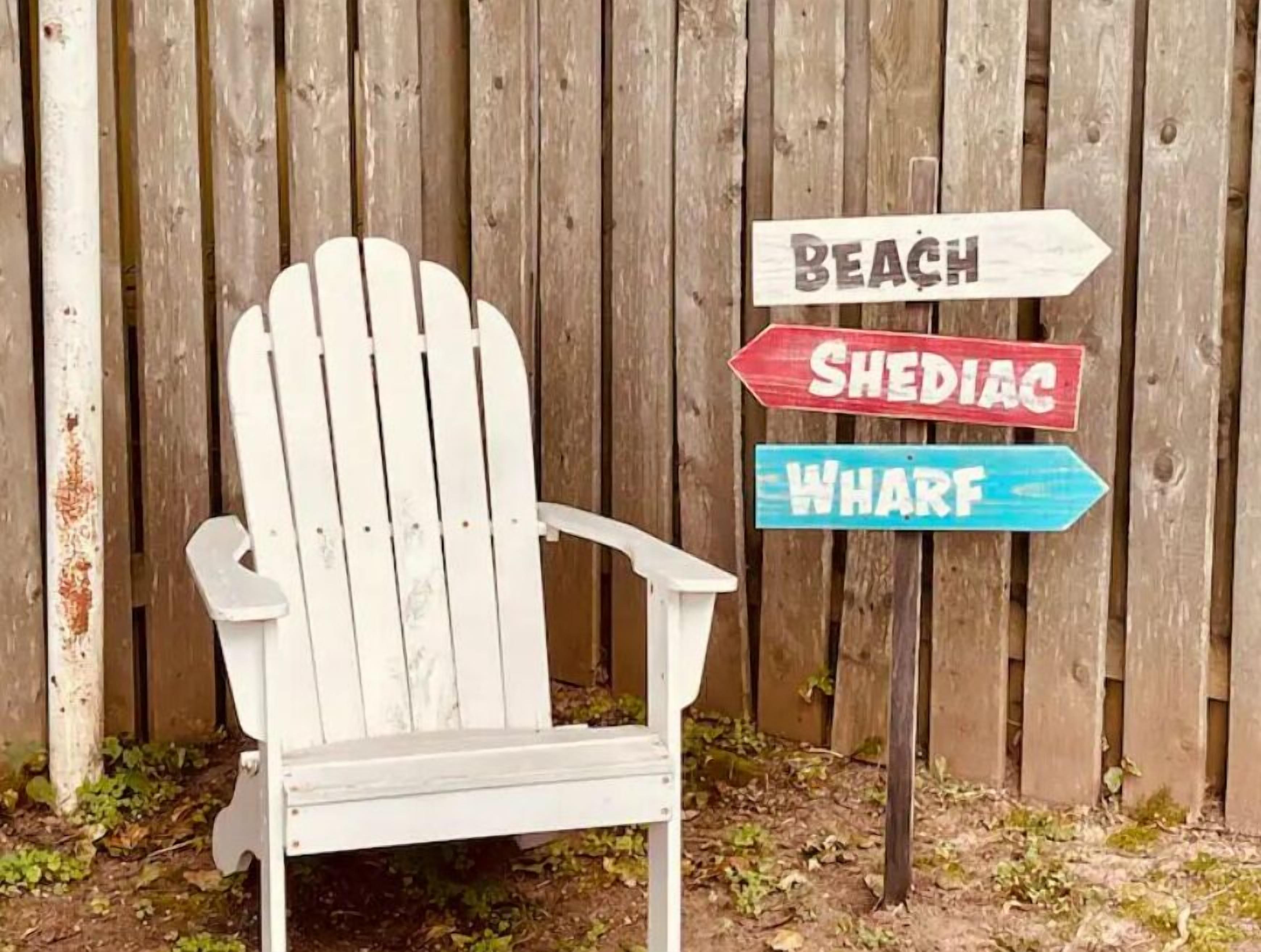 Ein weißer Adirondack-Stuhl vor einem Holzzaun mit Hinweisschildern, die zu 'Beach', 'Shediac' und 'Wharf' weisen.