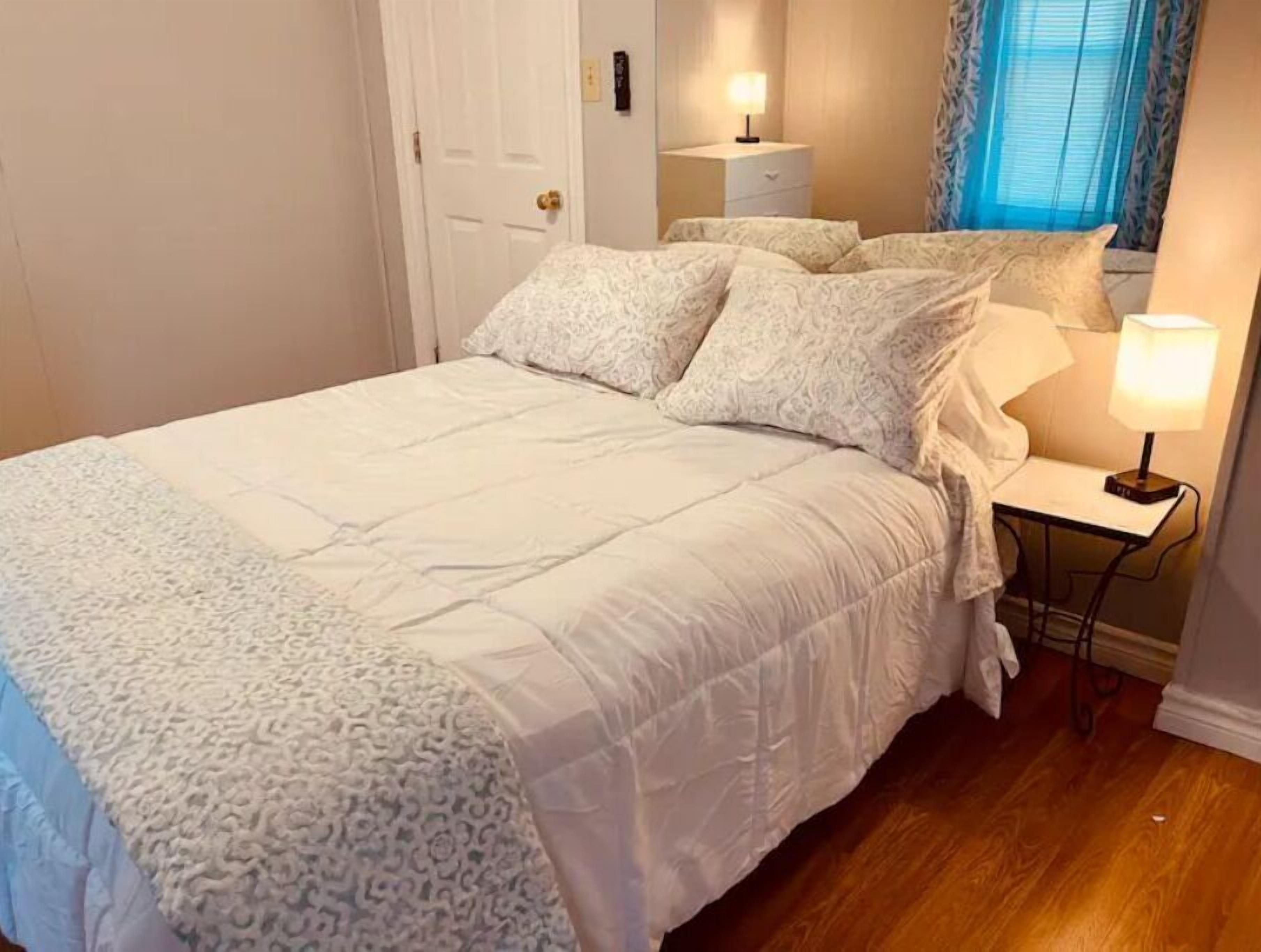 Una cama bien hecha con una colcha estampada y almohadas, una mesita de noche con una lámpara y una ventana con cortinas.
