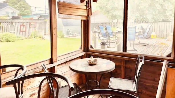 Une terrasse en bois avec une table ronde et des chaises, de grandes fenêtres et une vue sur un jardin clos, aménagé avec du mobilier d’extérieur.
