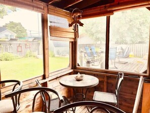 A wooden deck with a round table and chairs, large windows, and a view of a backyard with a fence and outdoor furniture. - On Pointe! (Pointe-du-Chêne)