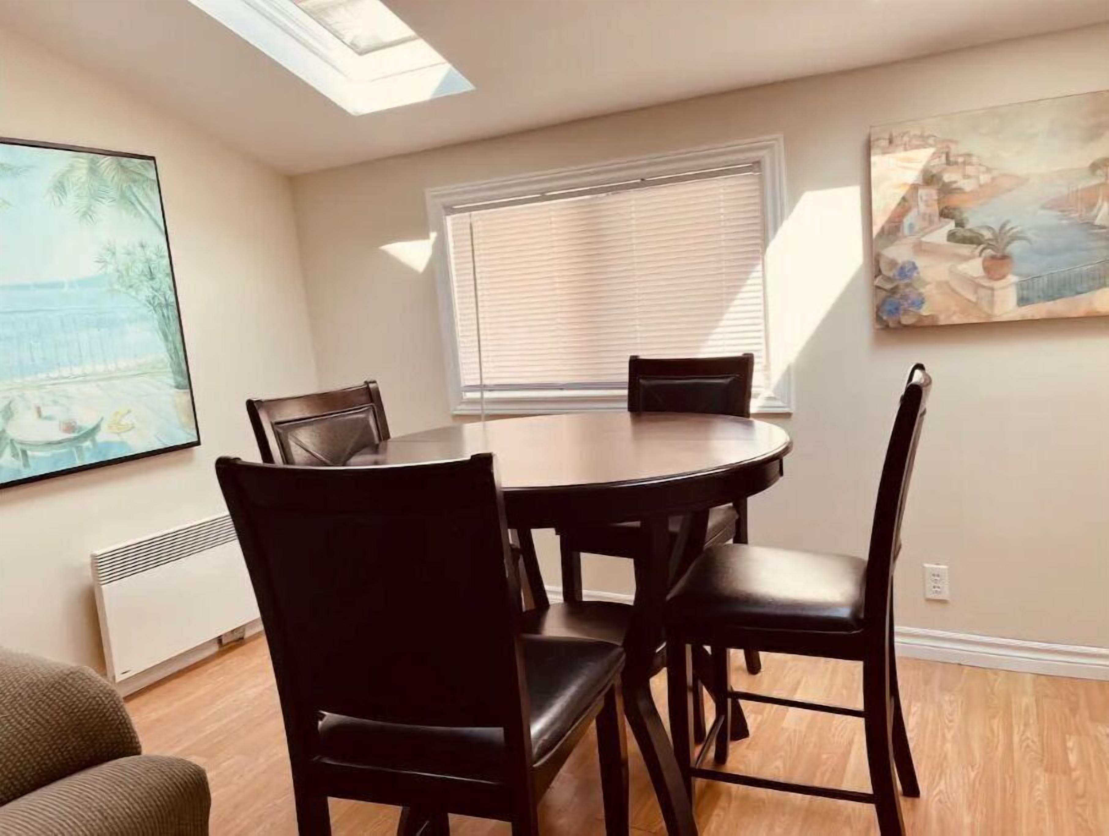A dining area with a round table and four chairs, a large window with blinds, a ceiling fan, and a painting on the wall.