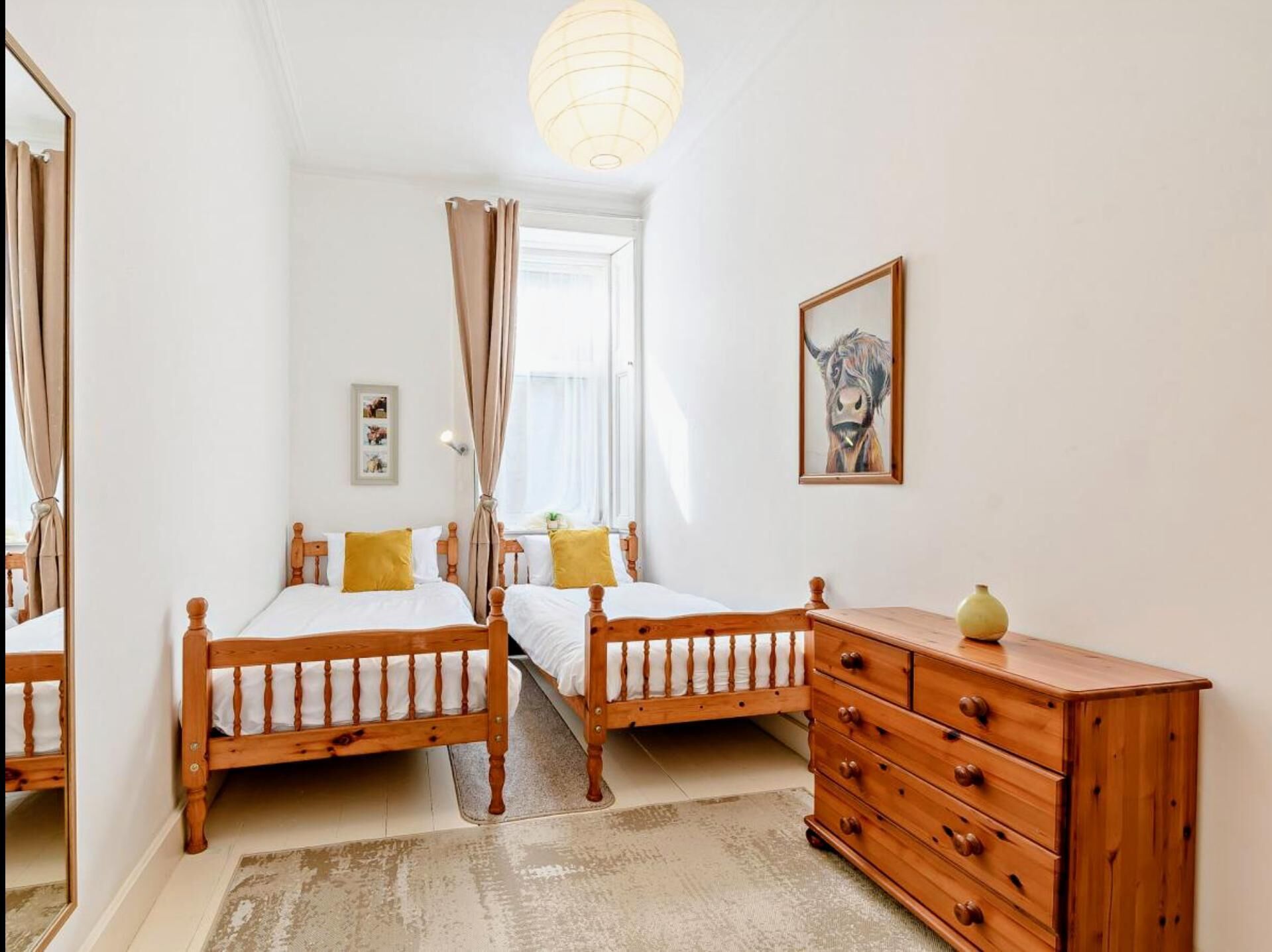A bedroom with two wooden beds, a wooden dresser, a mirror, a framed picture, and a large window with curtains.