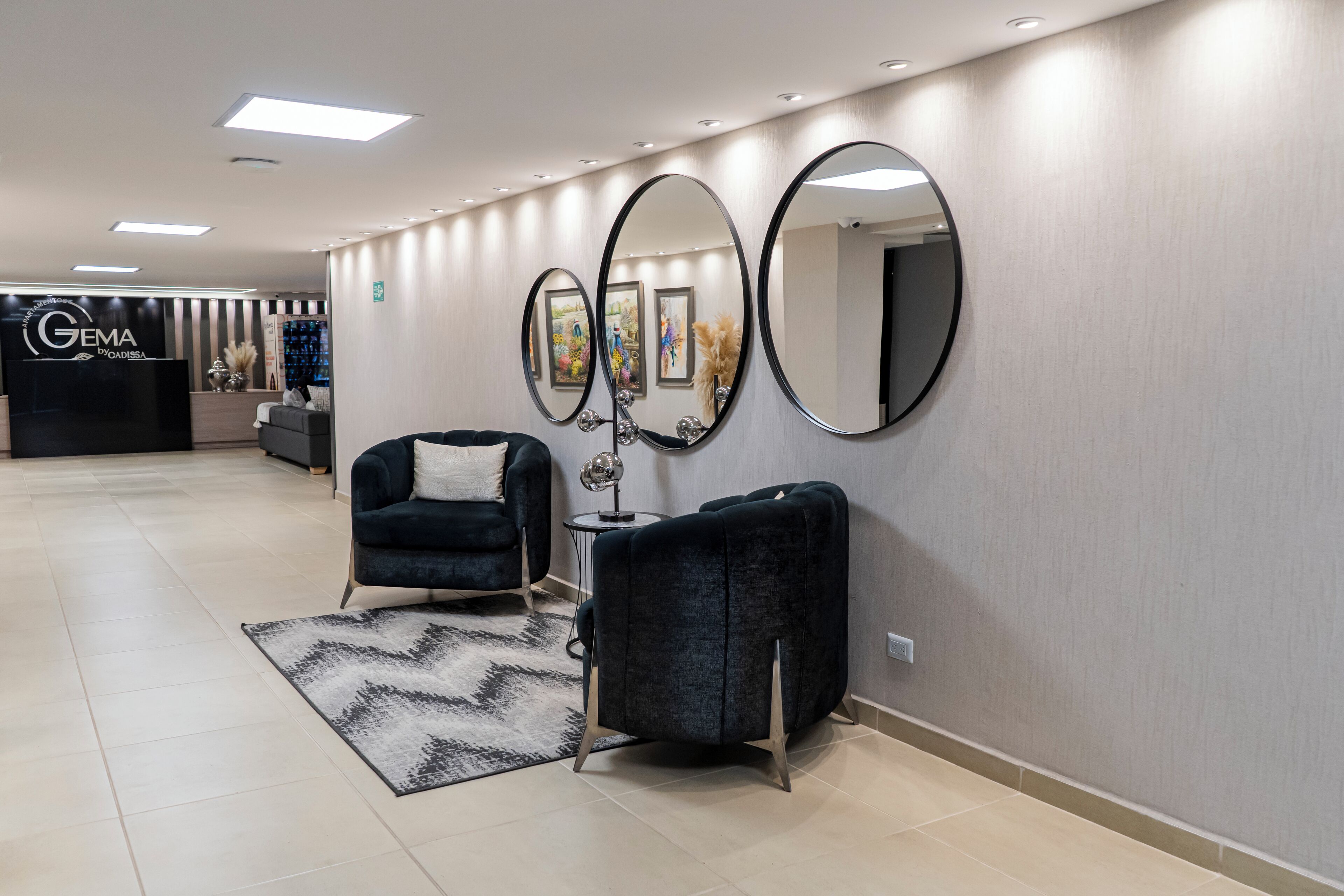 Una hall d'hotel moderna con una reception, due poltrone, uno specchio rotondo e un tappeto decorato.